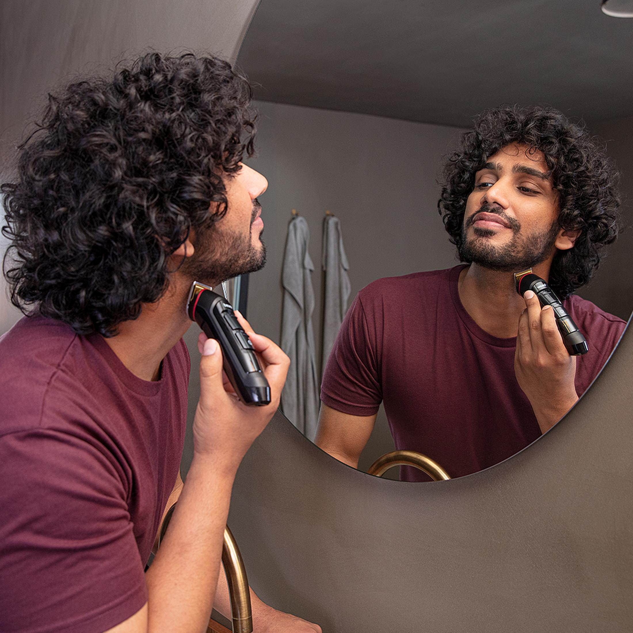 Man met krullend haar trimt baard in spiegel met trimmer. Donker shirt.