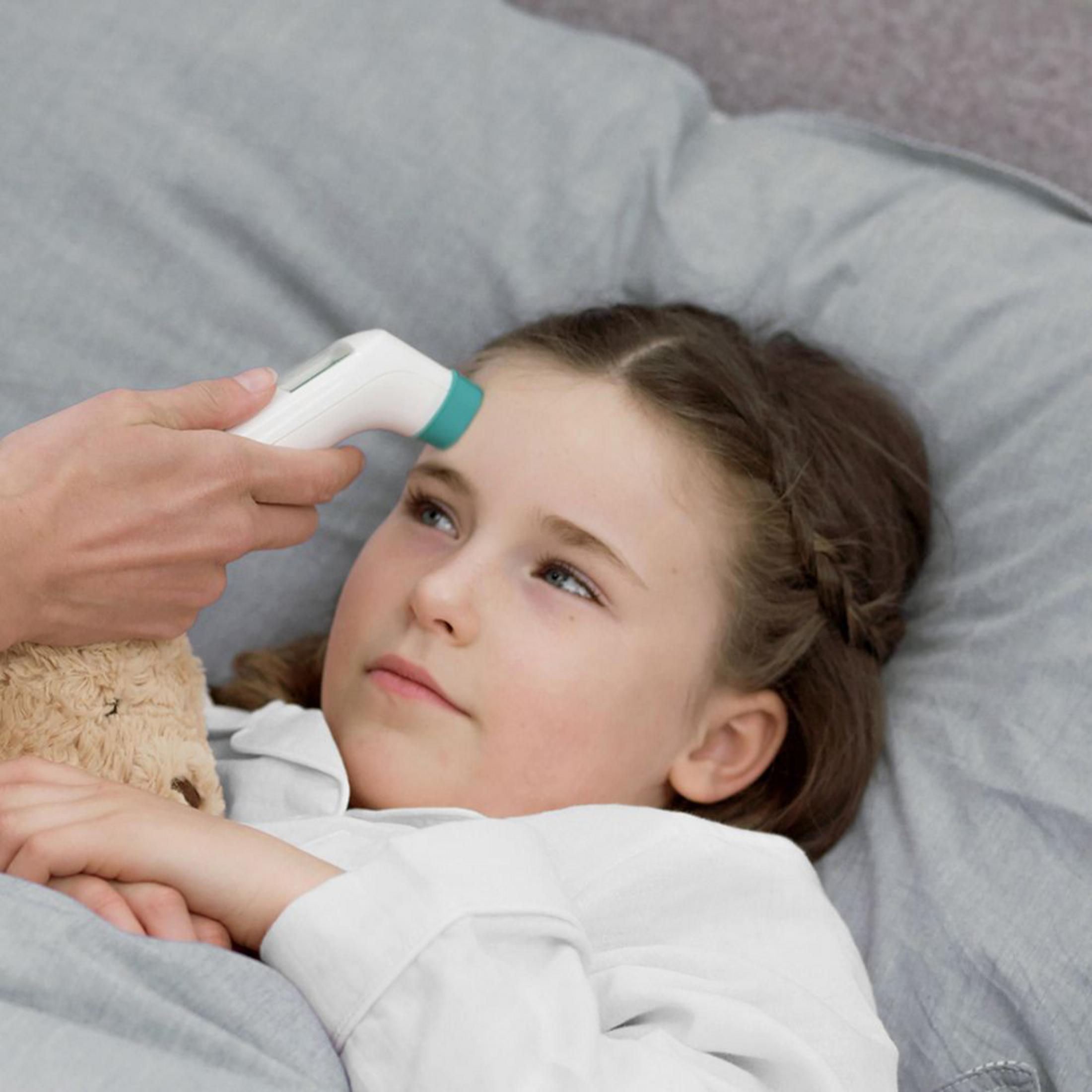 Termometr przyłożony do czoła dziewczynki leżącej na szarym łóżku.