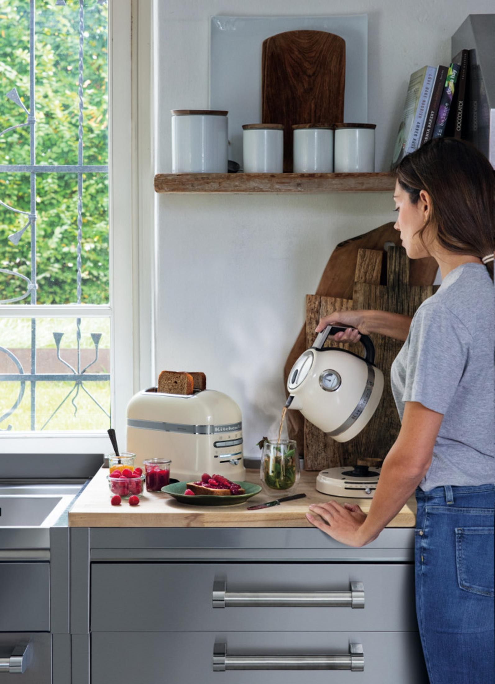 Kobieta nalewa wodę z czajnika do szklanki. Na stole leżą tosty i jagody.