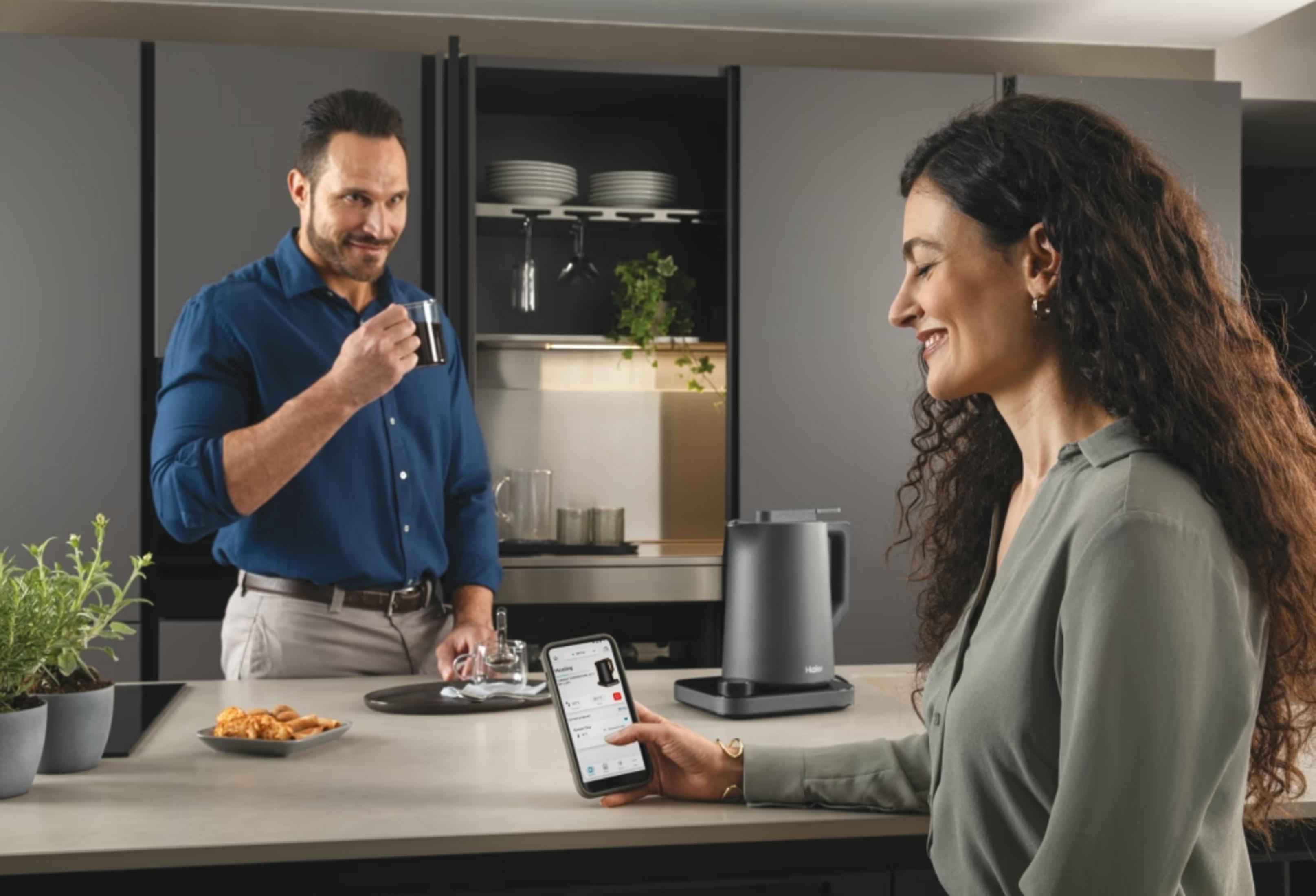 Mężczyzna pije kawę, kobieta używa smartfona obok czajnika.