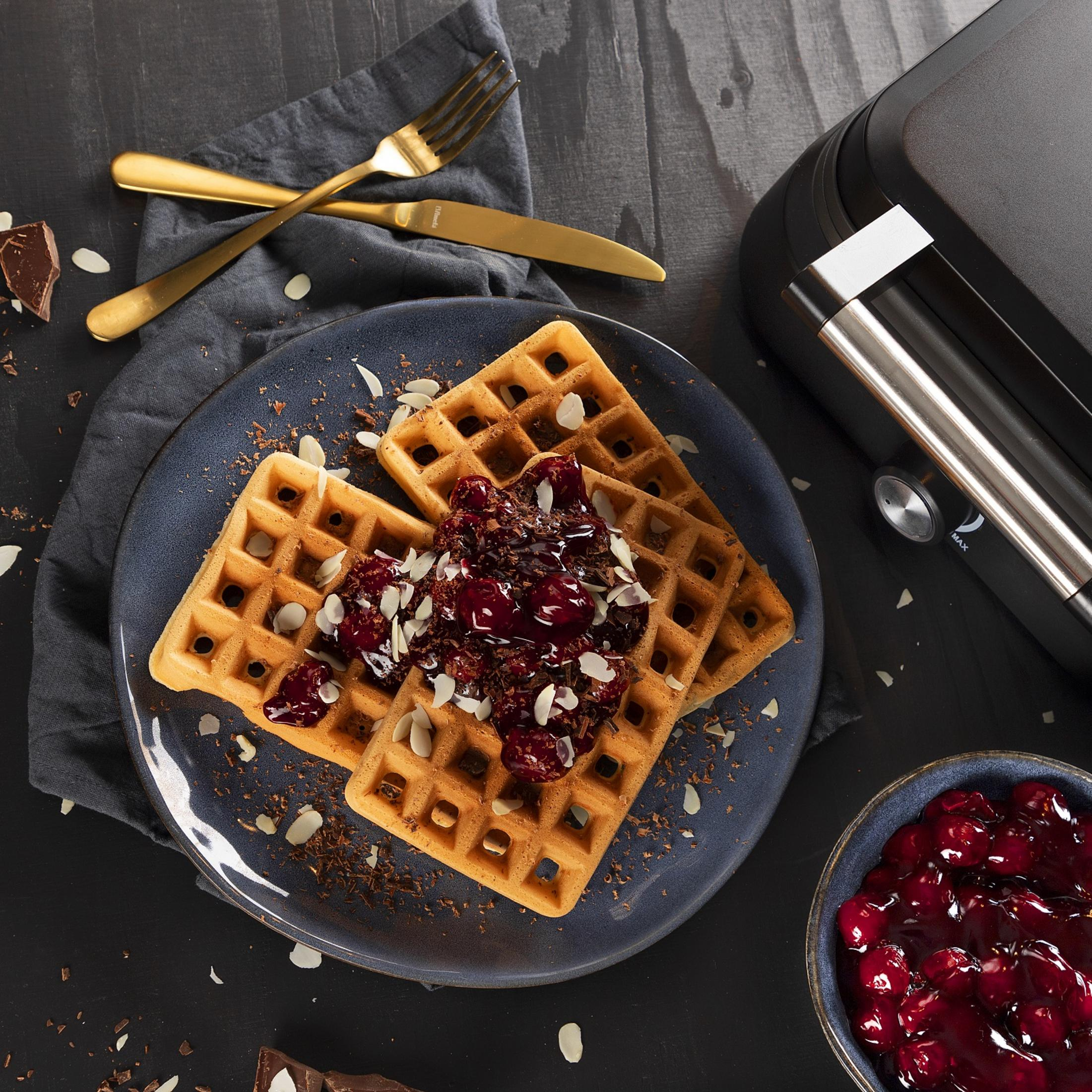 Dwa gofry z polewą wiśniową, wiórkami czekolady i płatkami migdałów na niebieskim talerzu.