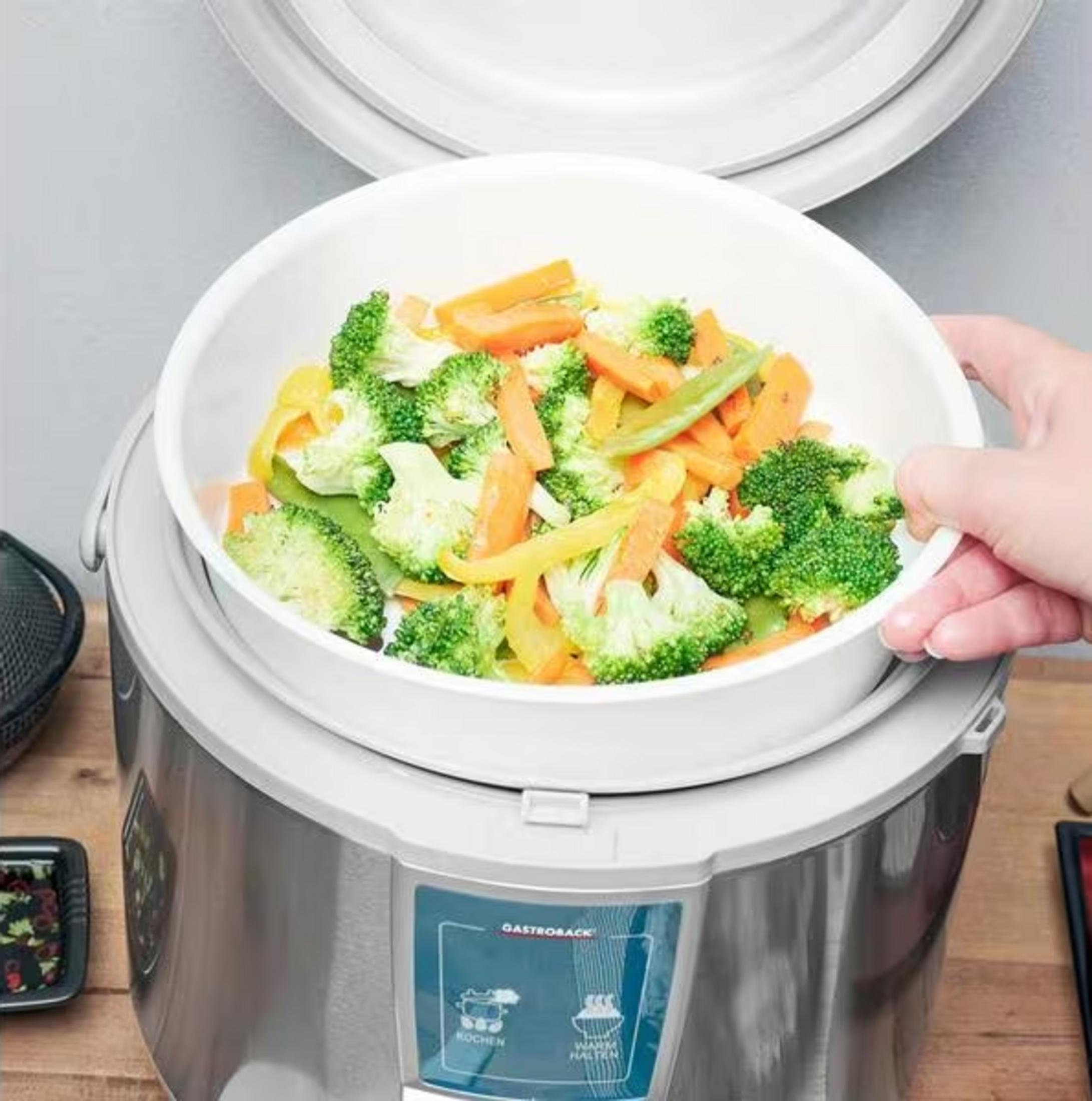 Una mano solleva una ciotola di broccoli, carote e peperoni al vapore da un cuocitore a vapore.