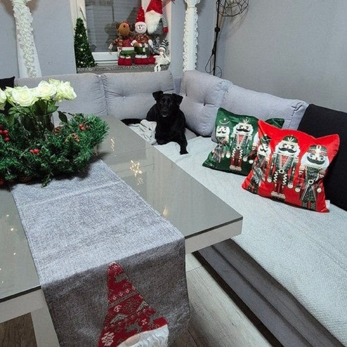 Un perro negro yace en un sofá junto a cojines navideños. Rosas blancas y ramas de pino en una mesa.