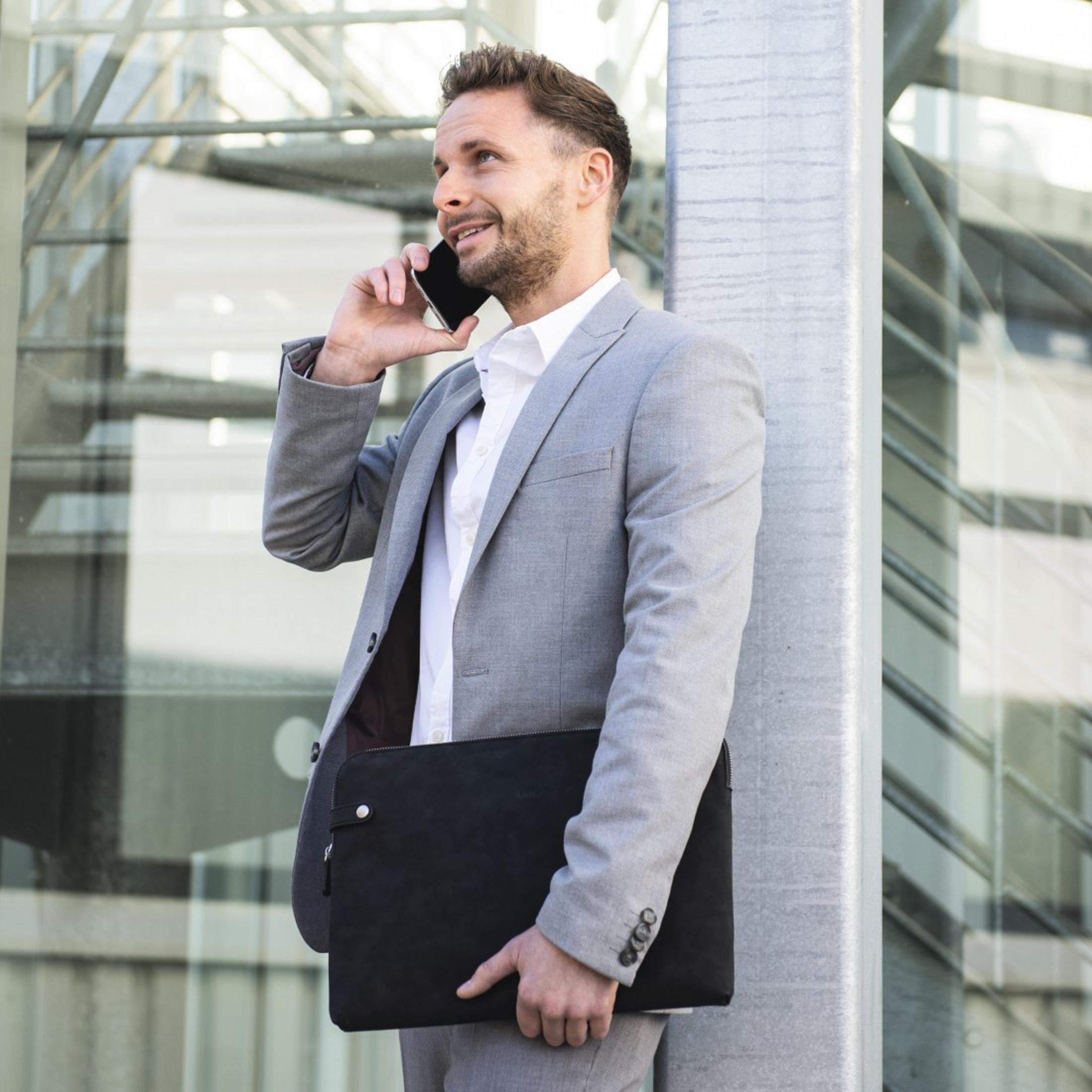 Mężczyzna w szarym garniturze rozmawia przez telefon, trzyma czarną torbę.
