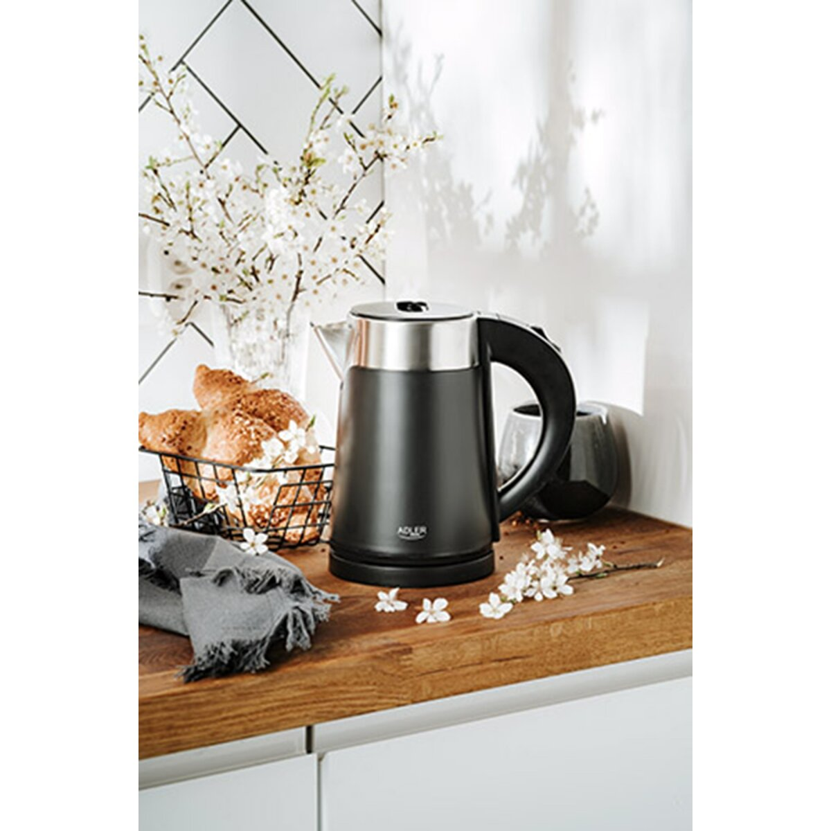 Czajnik elektryczny czarny z srebrną pokrywką obok koszyka z croissantami i białymi kwiatami.