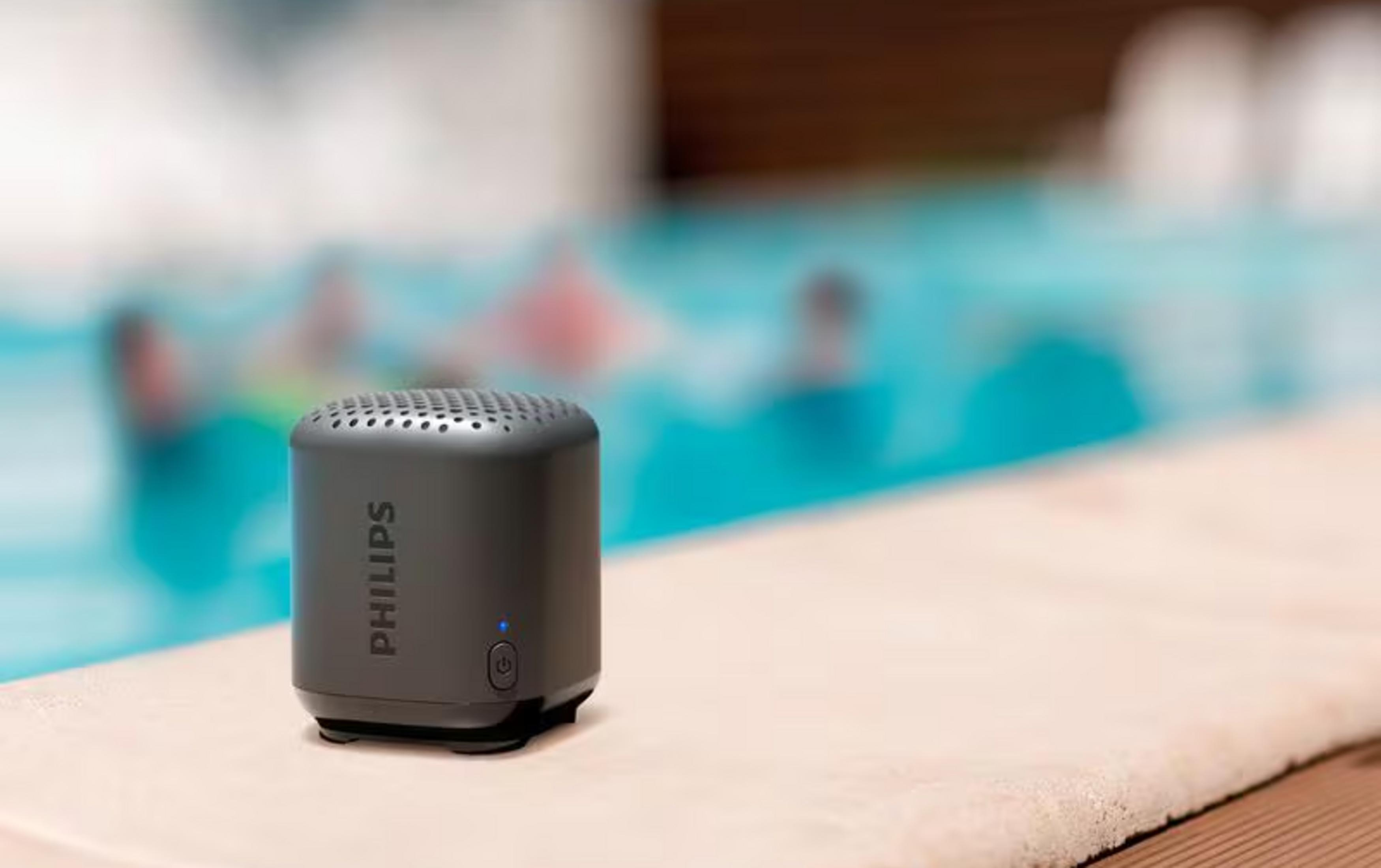 Szary głośnik Philips przy basenie z pływającymi ludźmi w tle.