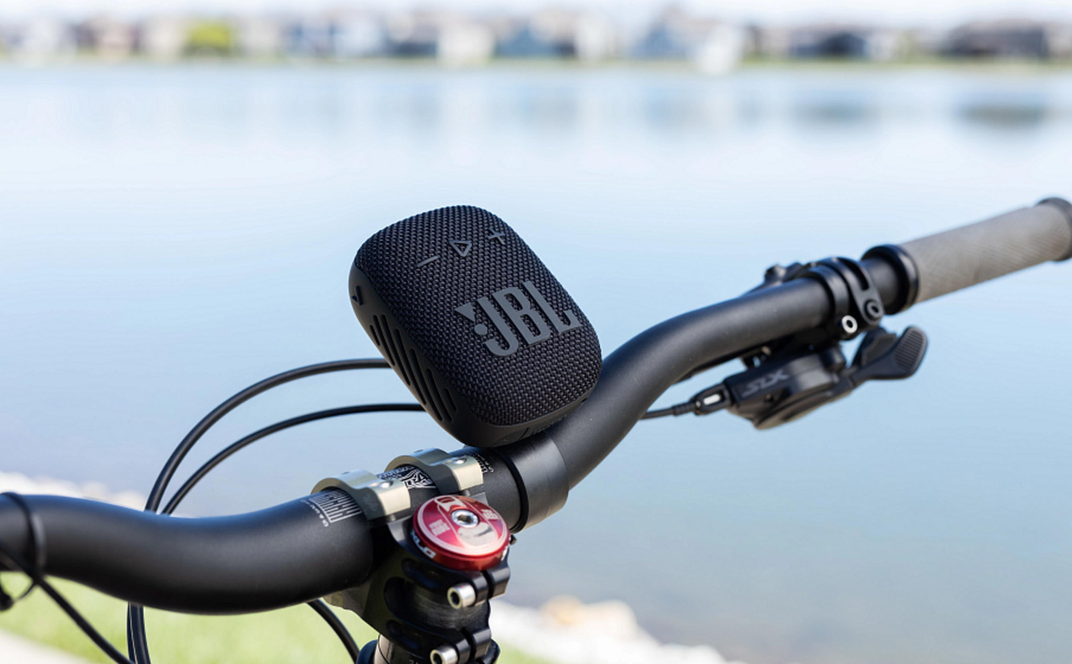 Schwarzer JBL-Lautsprecher am Lenker eines Fahrrads, mit See und Häusern im Hintergrund.