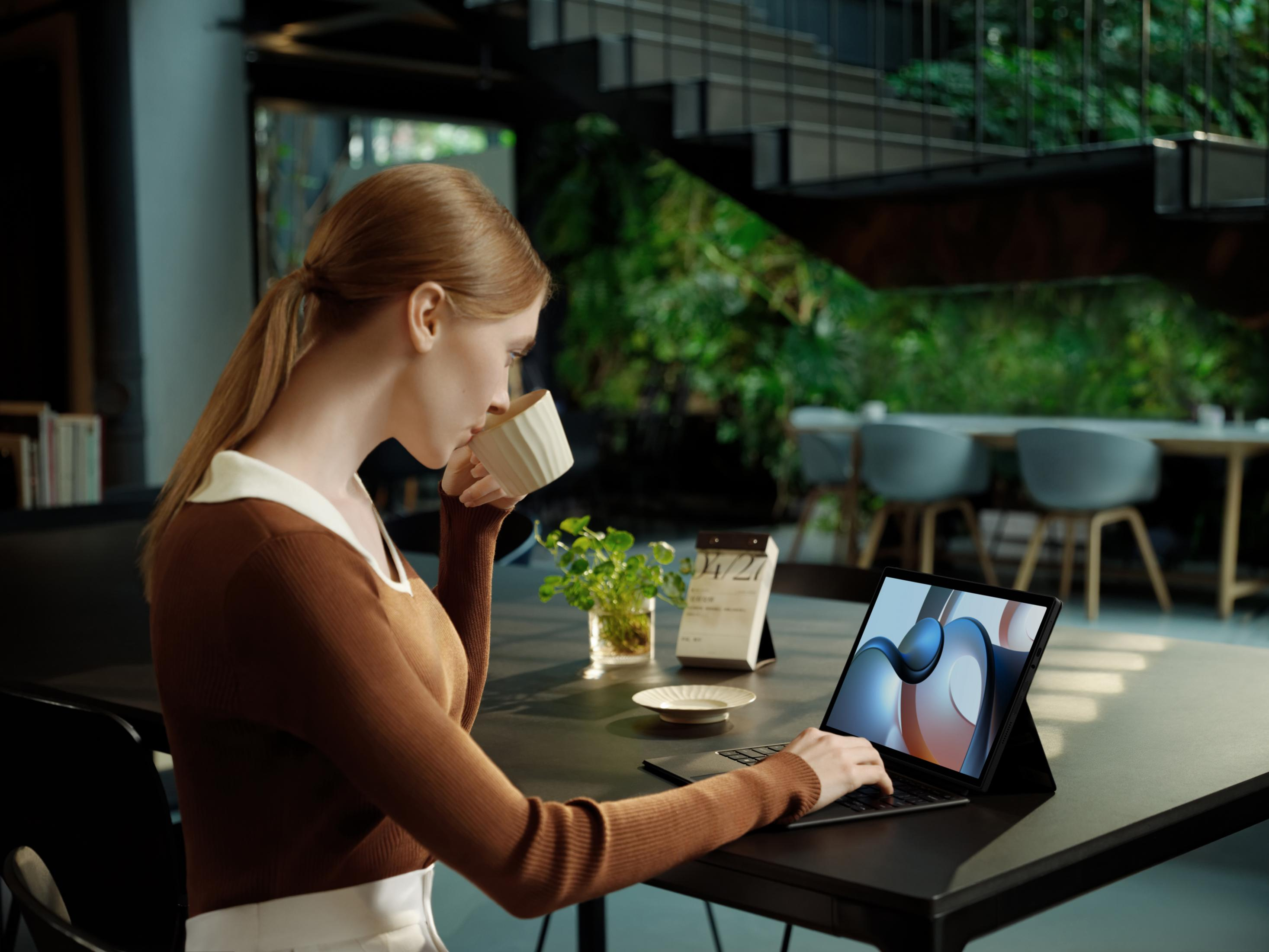 Een vrouw drinkt koffie en werkt op een laptop in een modern kantoor met planten.