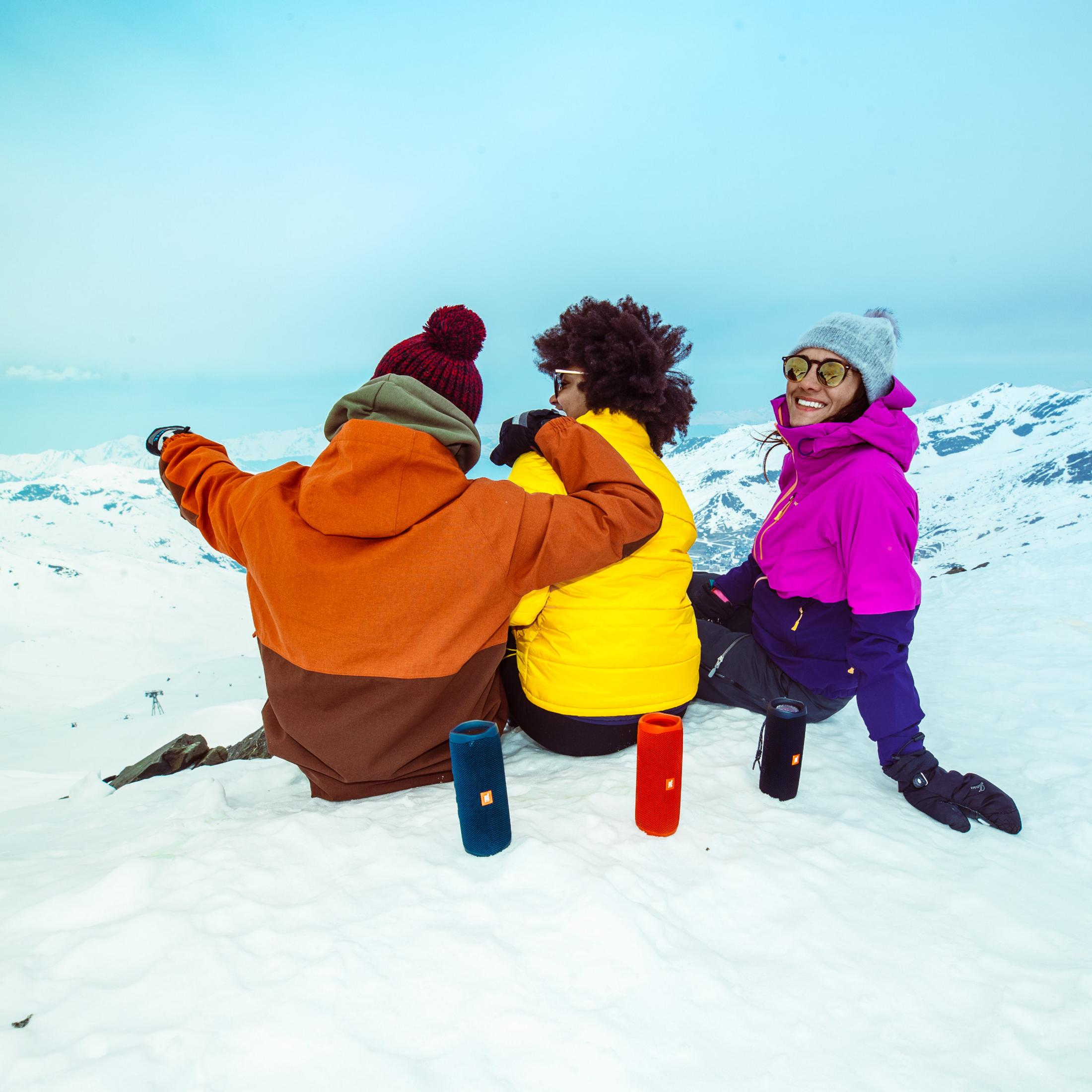 Drei Freunde sitzen im Schnee mit JBL-Lautsprechern, Berge im Hintergrund.