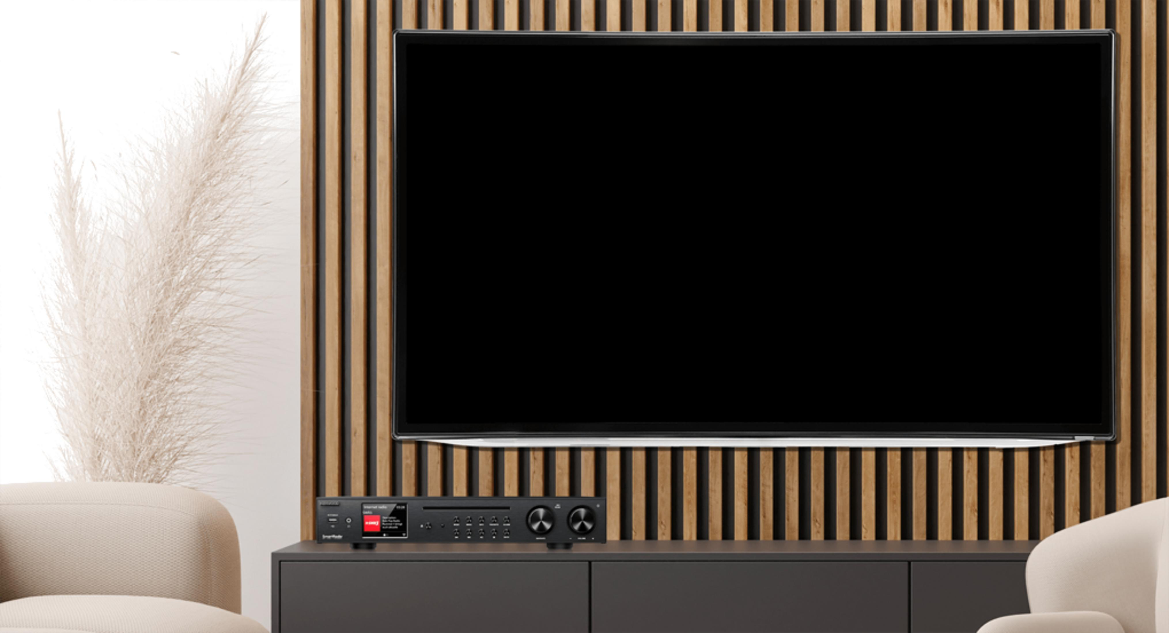 A black receiver sits on a dark cabinet in front of a flat-screen TV.