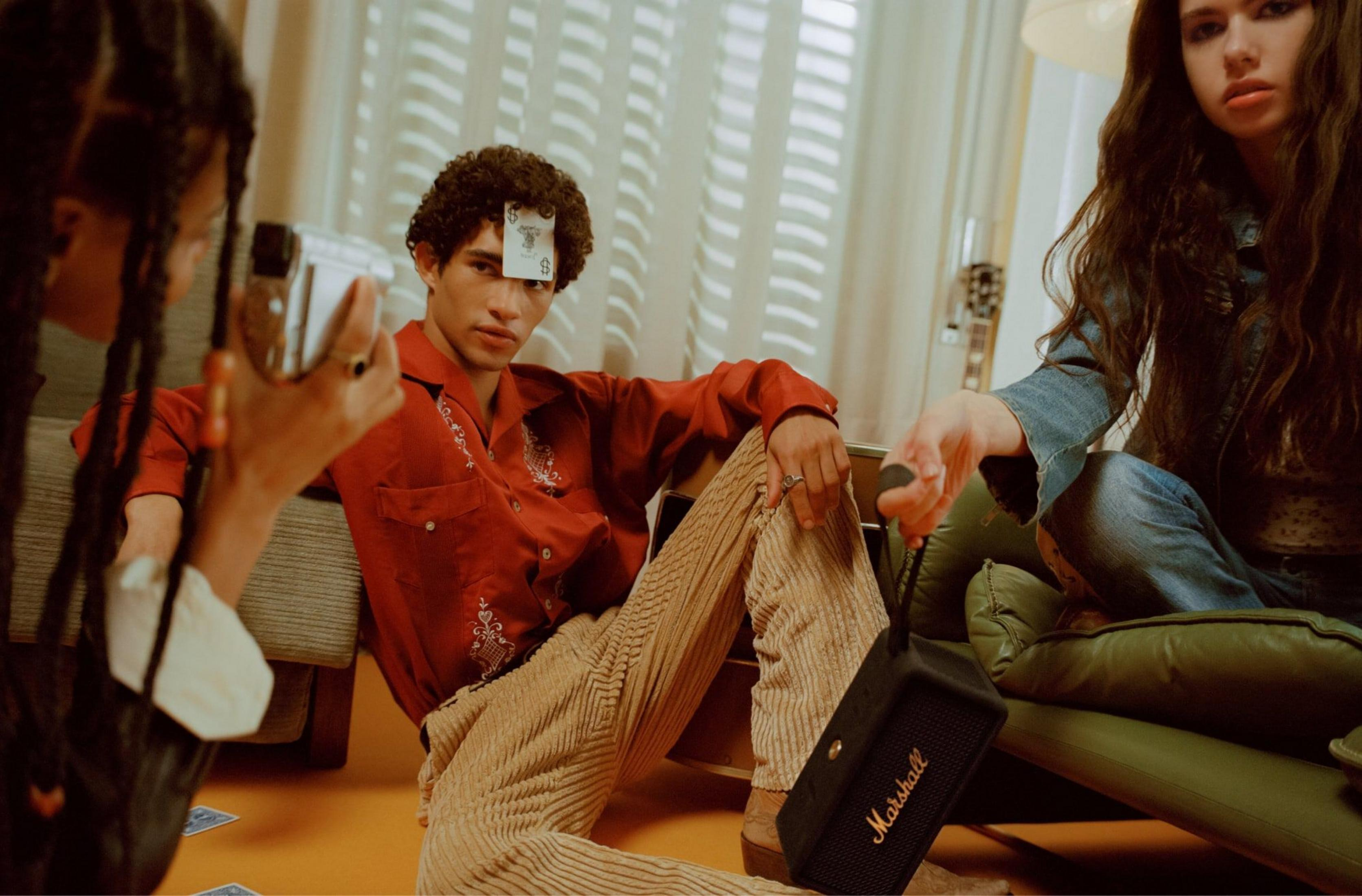 Drei junge Leute sitzen auf dem Boden, eine Karte steckt auf der Stirn eines Mannes.