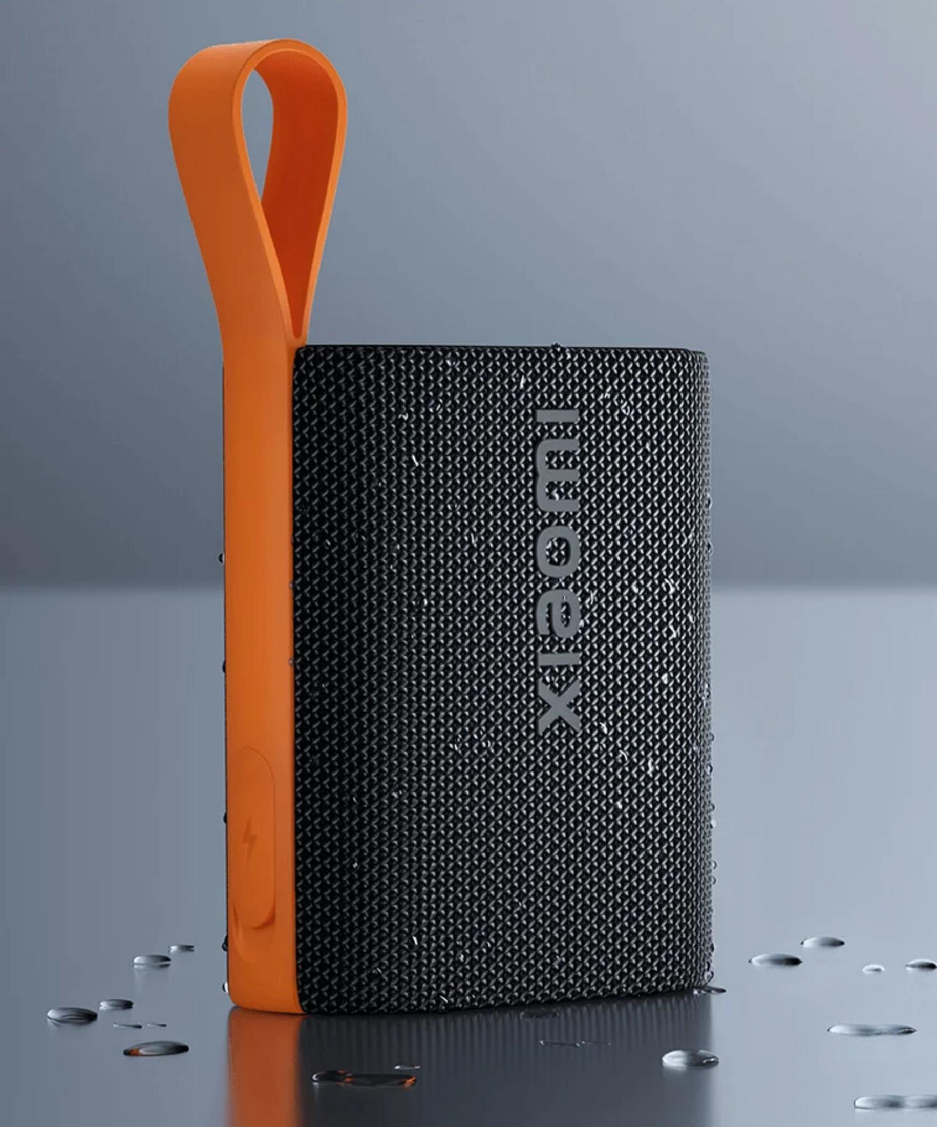 Czarny głośnik Xiaomi z pomarańczowym uchwytem, otoczony kroplami wody.