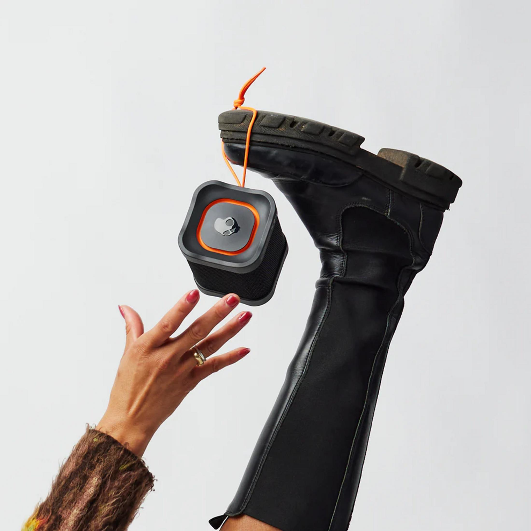 Ein schwarzer Stiefel hält einen kleinen, grauen Lautsprecher mit orangem Rand.