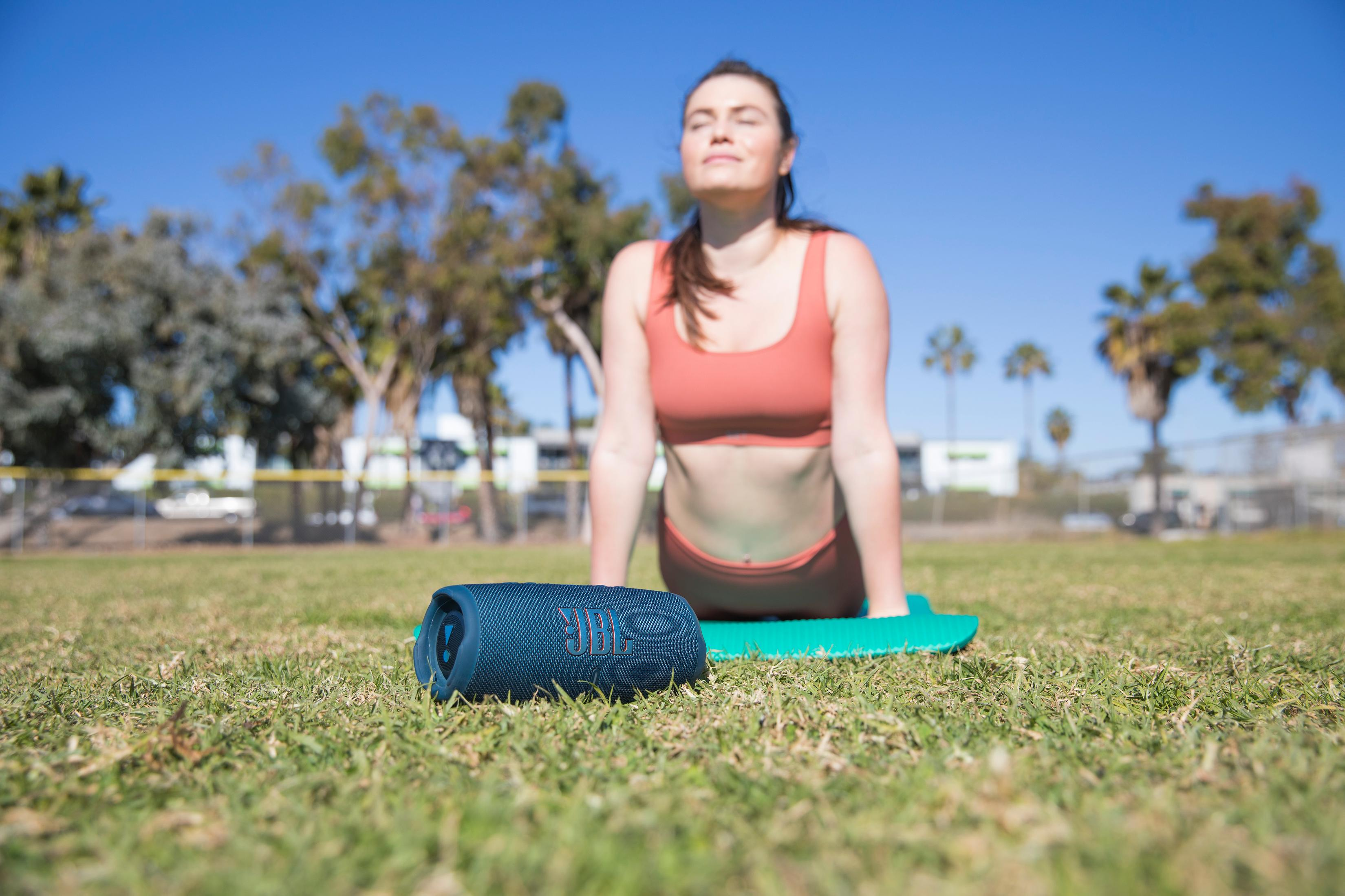 Kobieta uprawia jogę na macie na zewnątrz obok niebieskiego głośnika JBL.