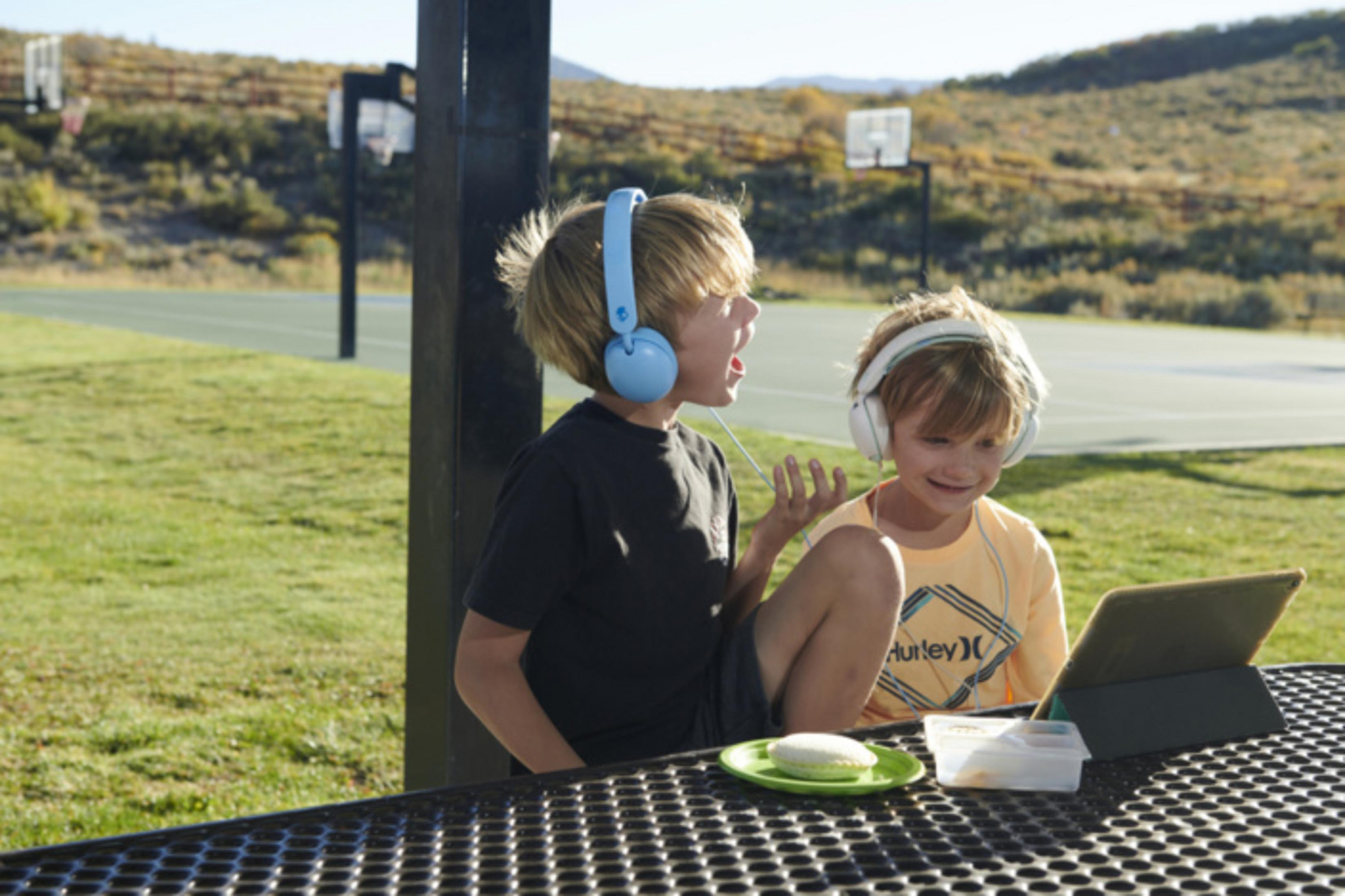 Dwóch chłopców w słuchawkach siedzi na zewnątrz przy stole, patrząc na tablet.