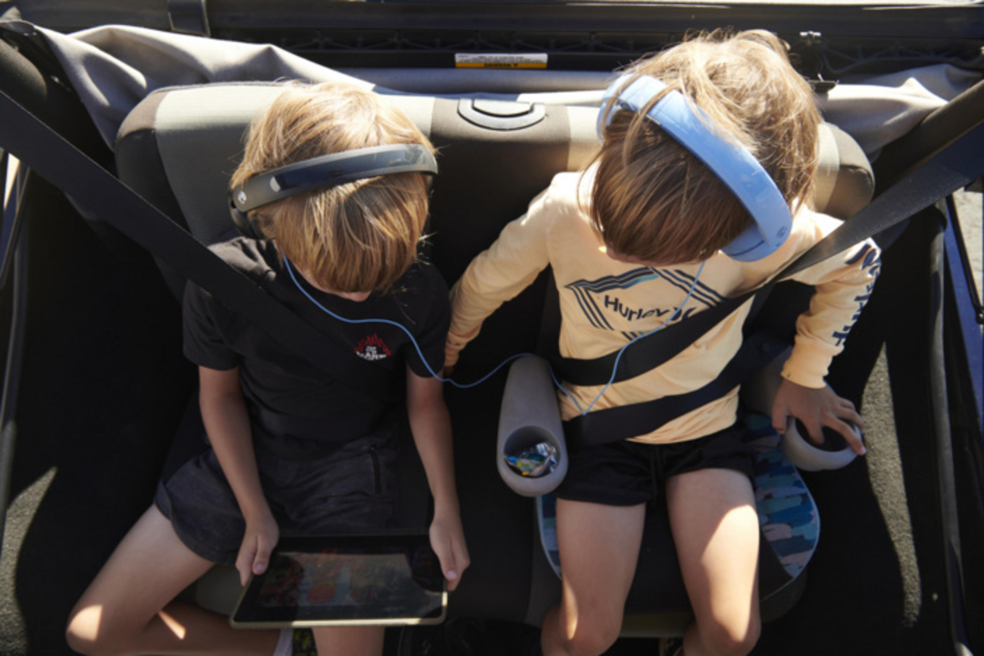 Dwoje dzieci w słuchawkach i z tabletem siedzi zapiętych w samochodzie.