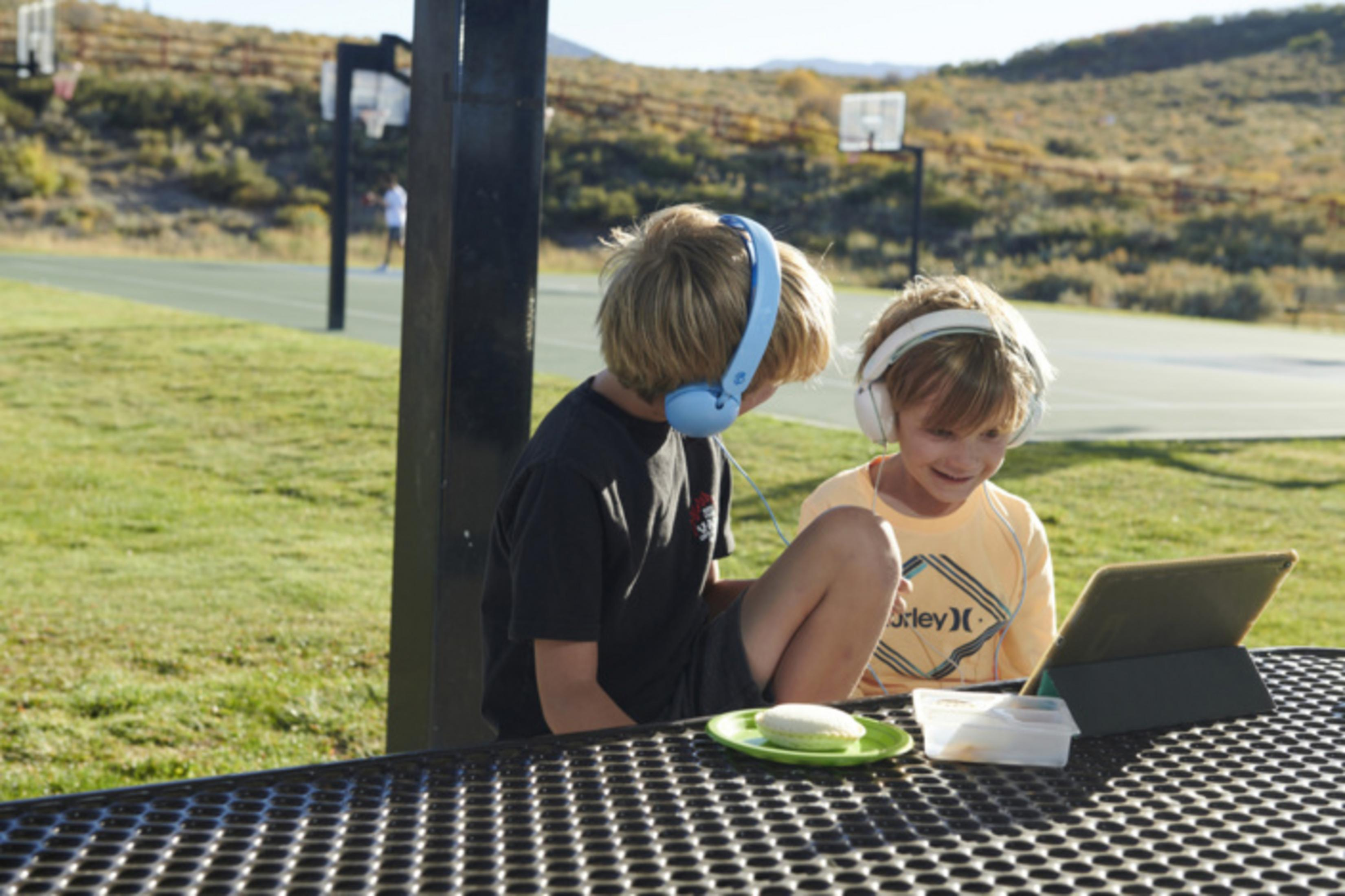 Dos niños con auriculares ven una tableta en una mesa de picnic al aire libre cerca de una cancha de baloncesto.