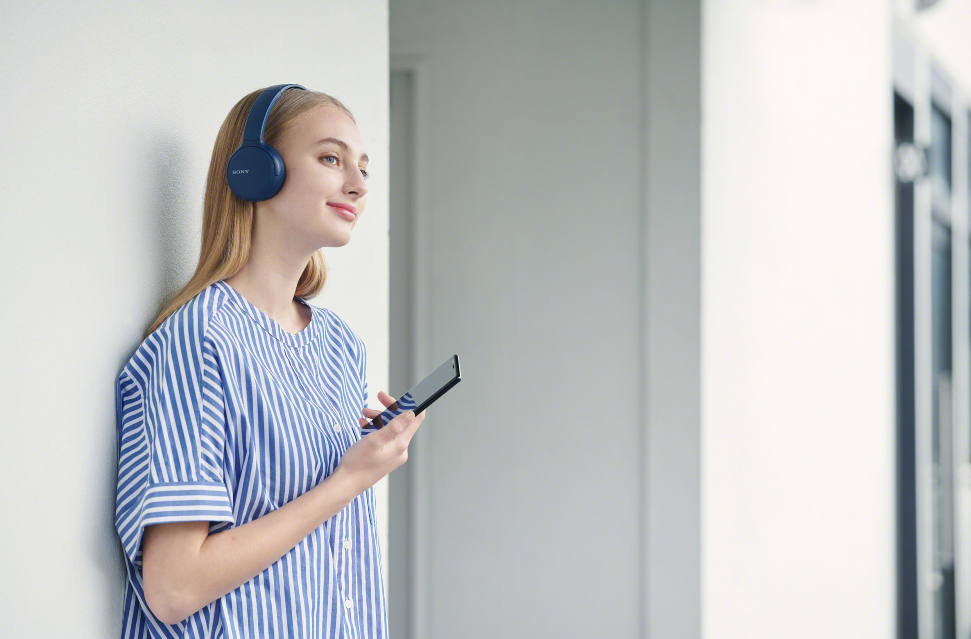 Una joven con auriculares se apoya en una pared, sosteniendo un smartphone.