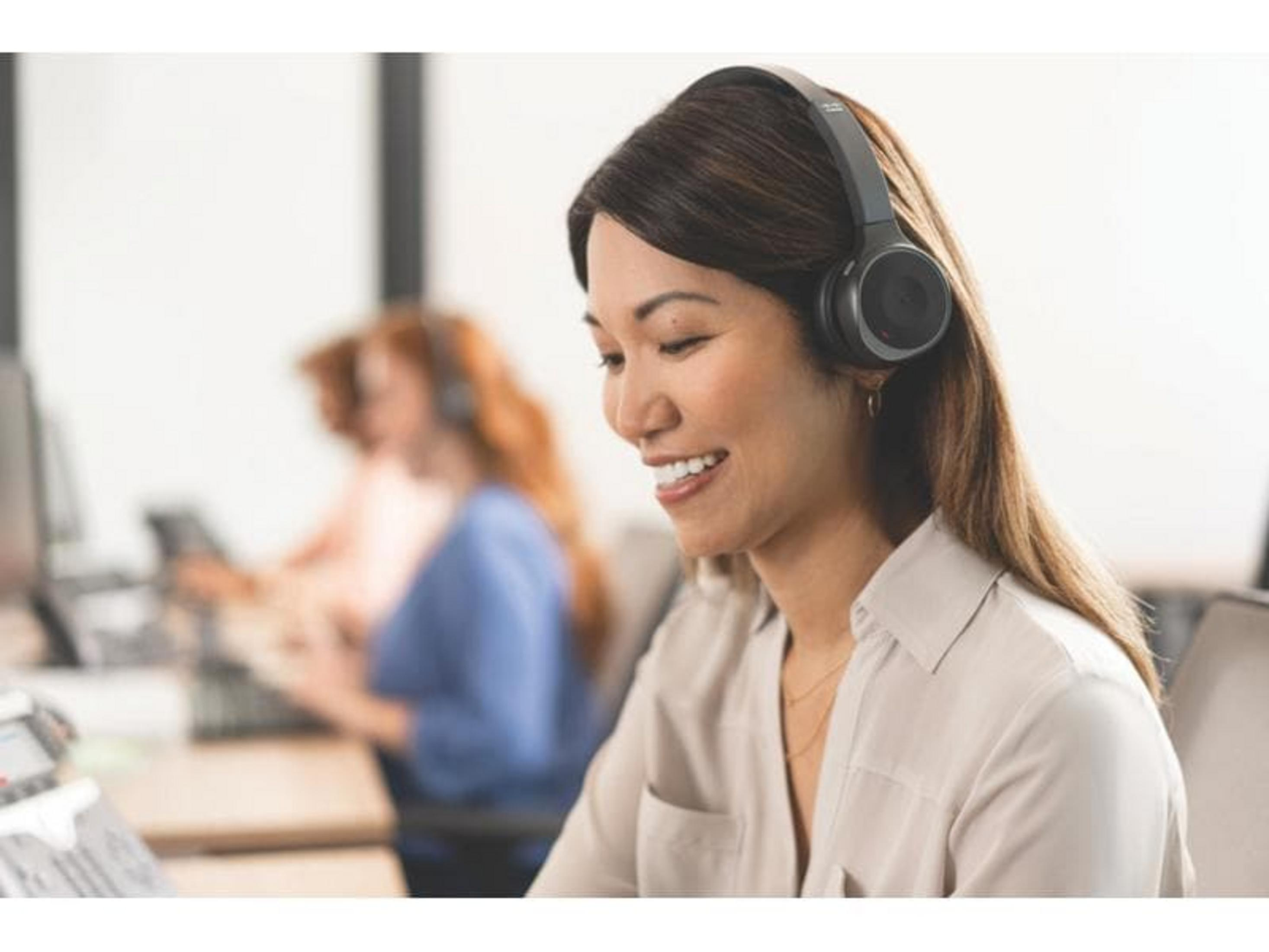 Kobieta w słuchawkach uśmiecha się, pracując przy biurku. W tle inni koledzy.