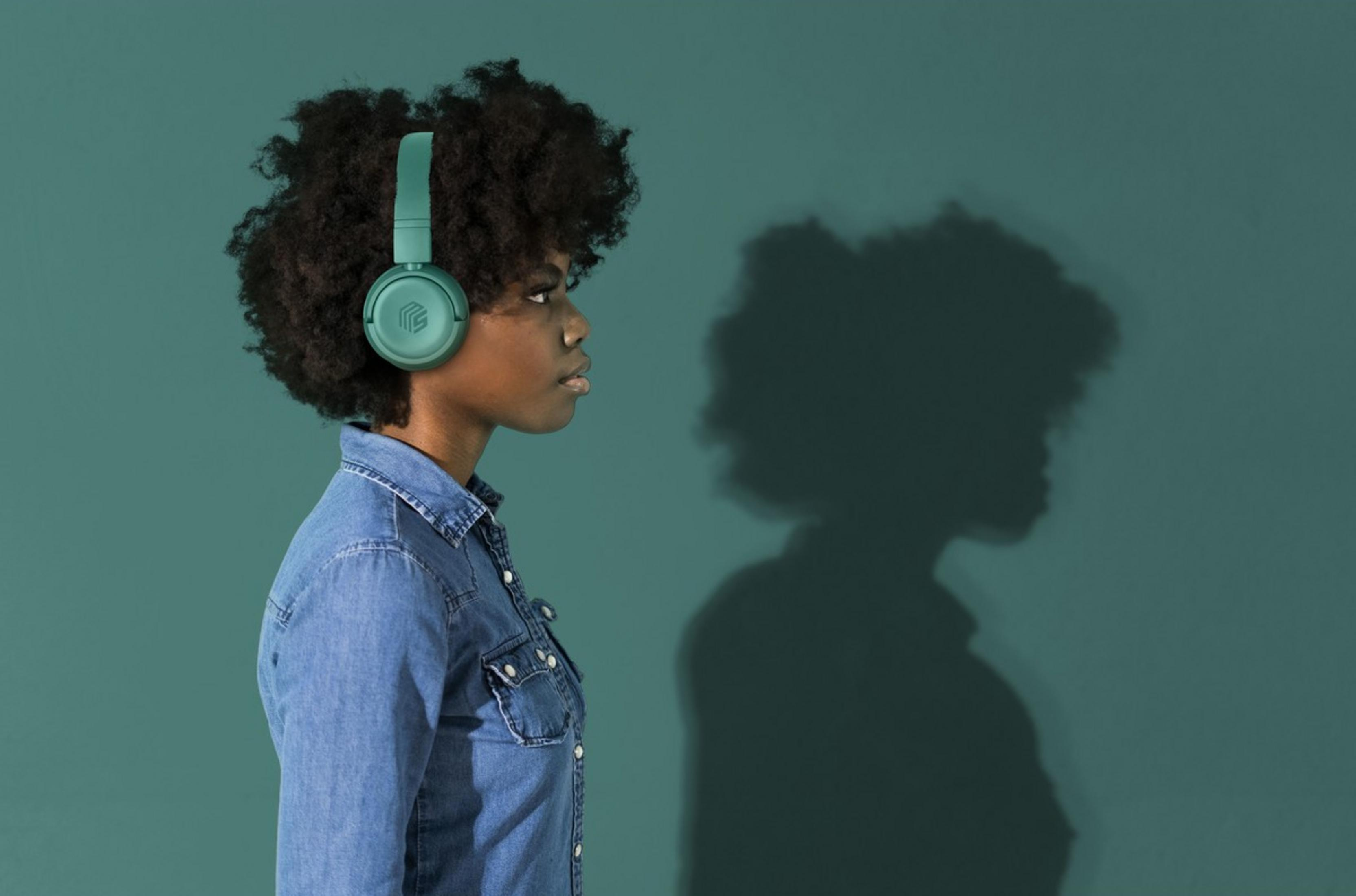 Eine Frau mit Afrohaaren trägt Kopfhörer und ein Jeanshemd vor einem teal Hintergrund.