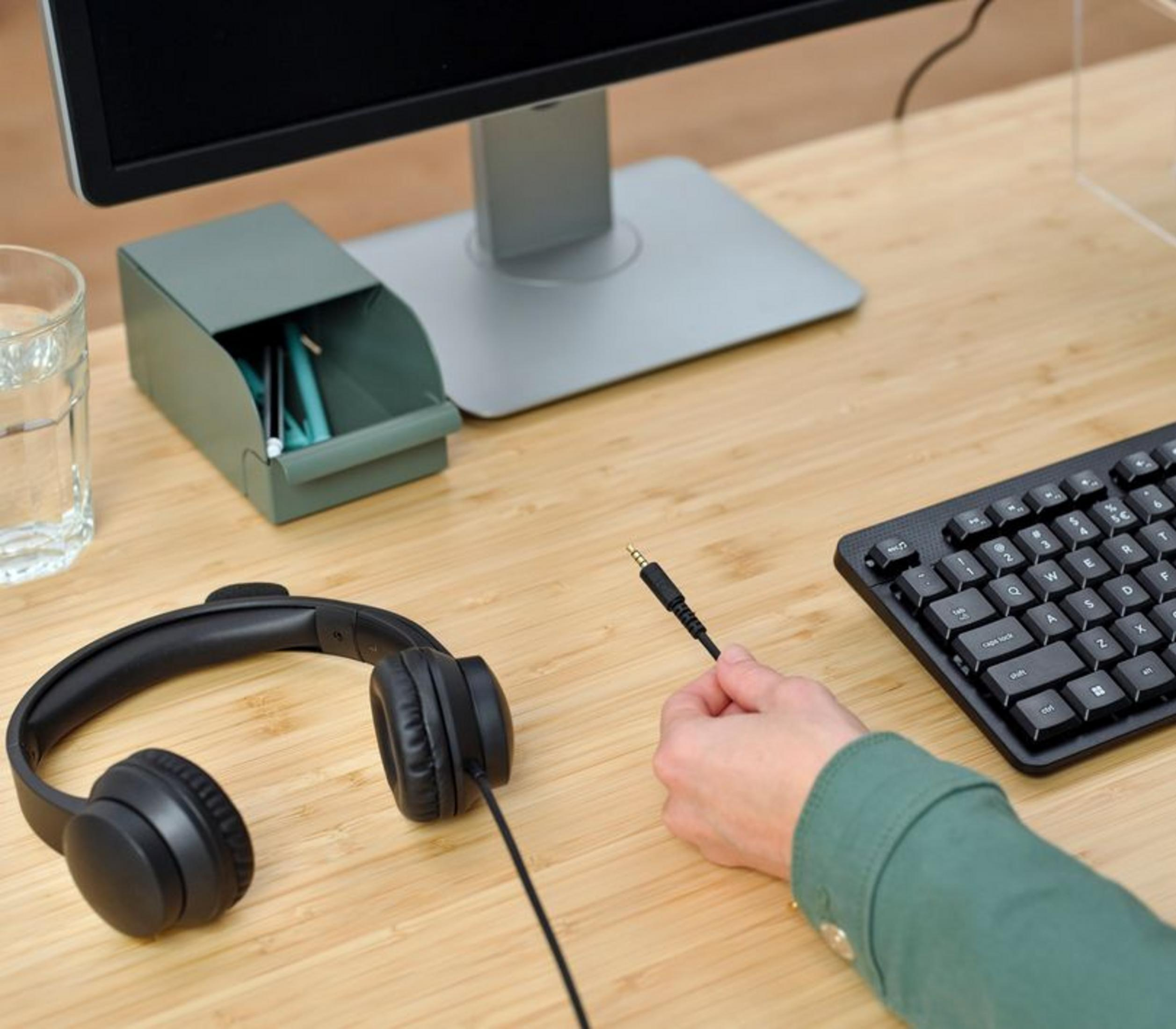 Słuchawki, klawiatura, monitor, pojemnik na długopisy i szklanka na drewnianym biurku.