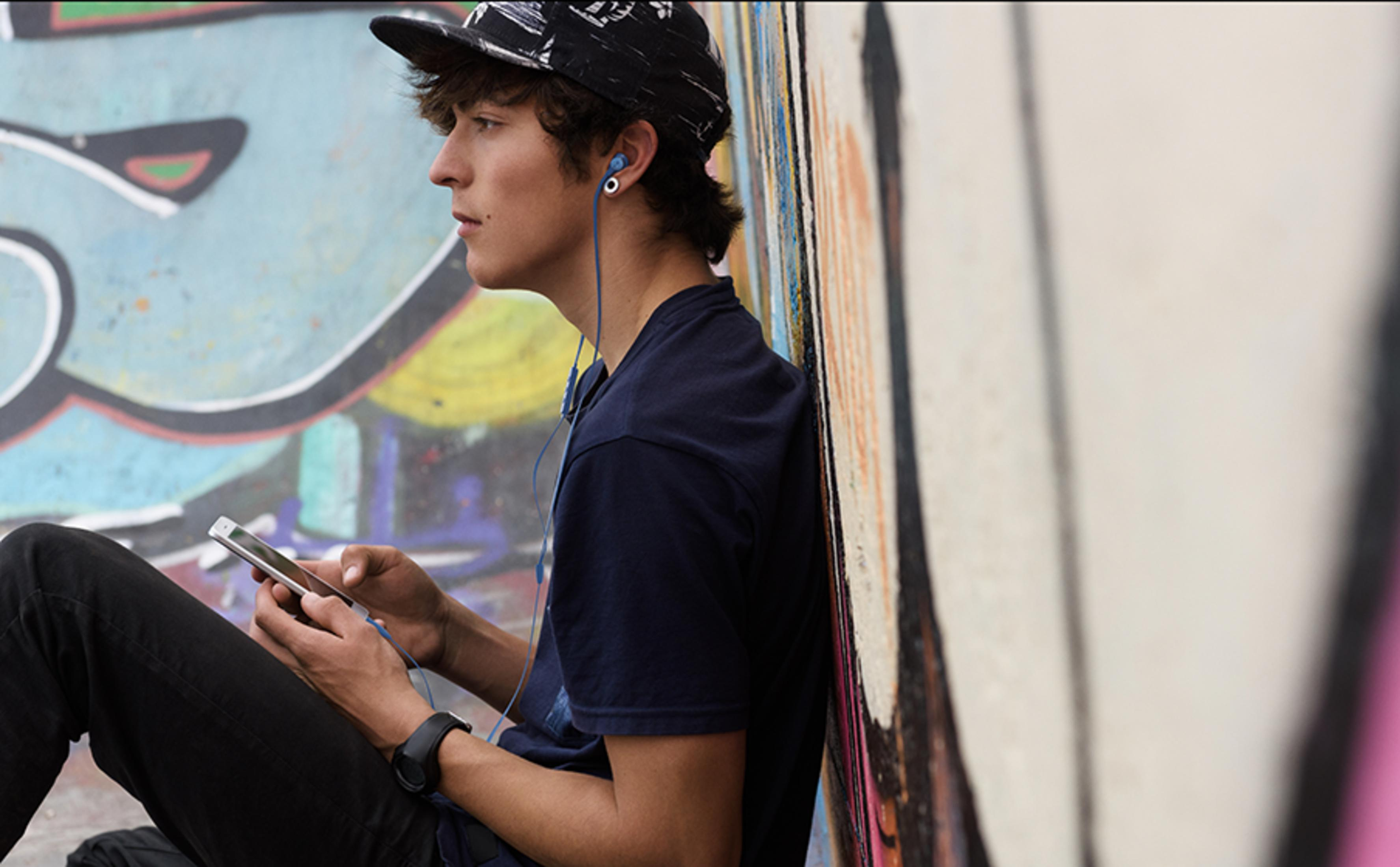 Młody mężczyzna ze słuchawkami i telefonem siedzi pod kolorowym murem z graffiti.