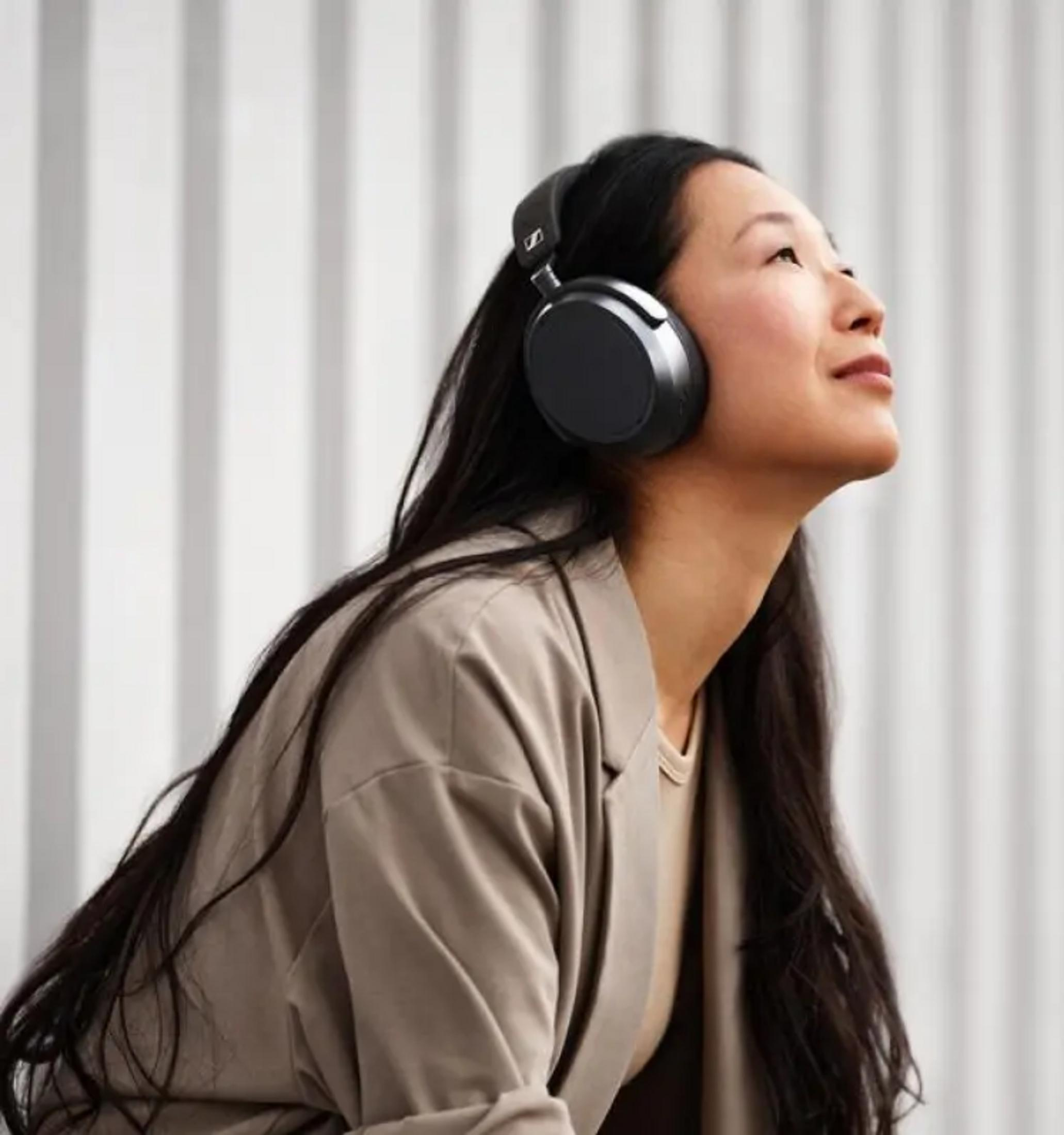 Kobieta w słuchawkach patrzy w górę, ubrana w beżową kurtkę. Pionowe tło.