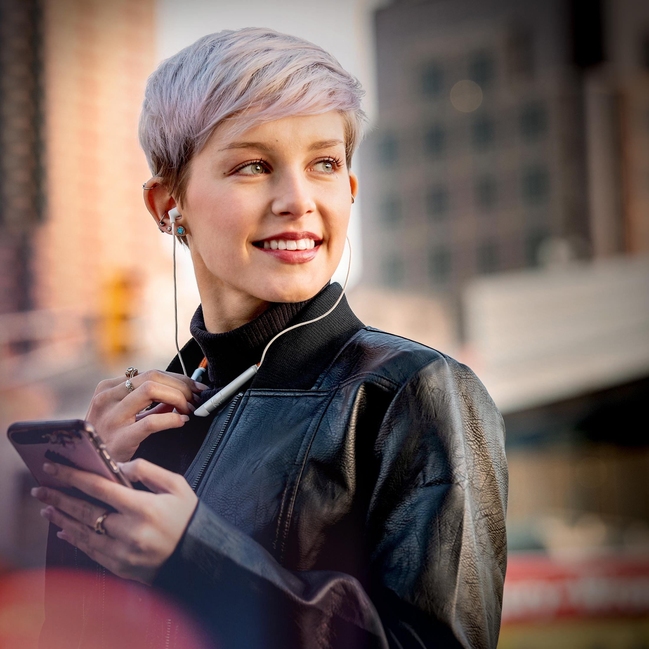 Eine junge Frau mit kurzen, hellen Haaren hört Musik über Kopfhörer und hält ein Smartphone.
