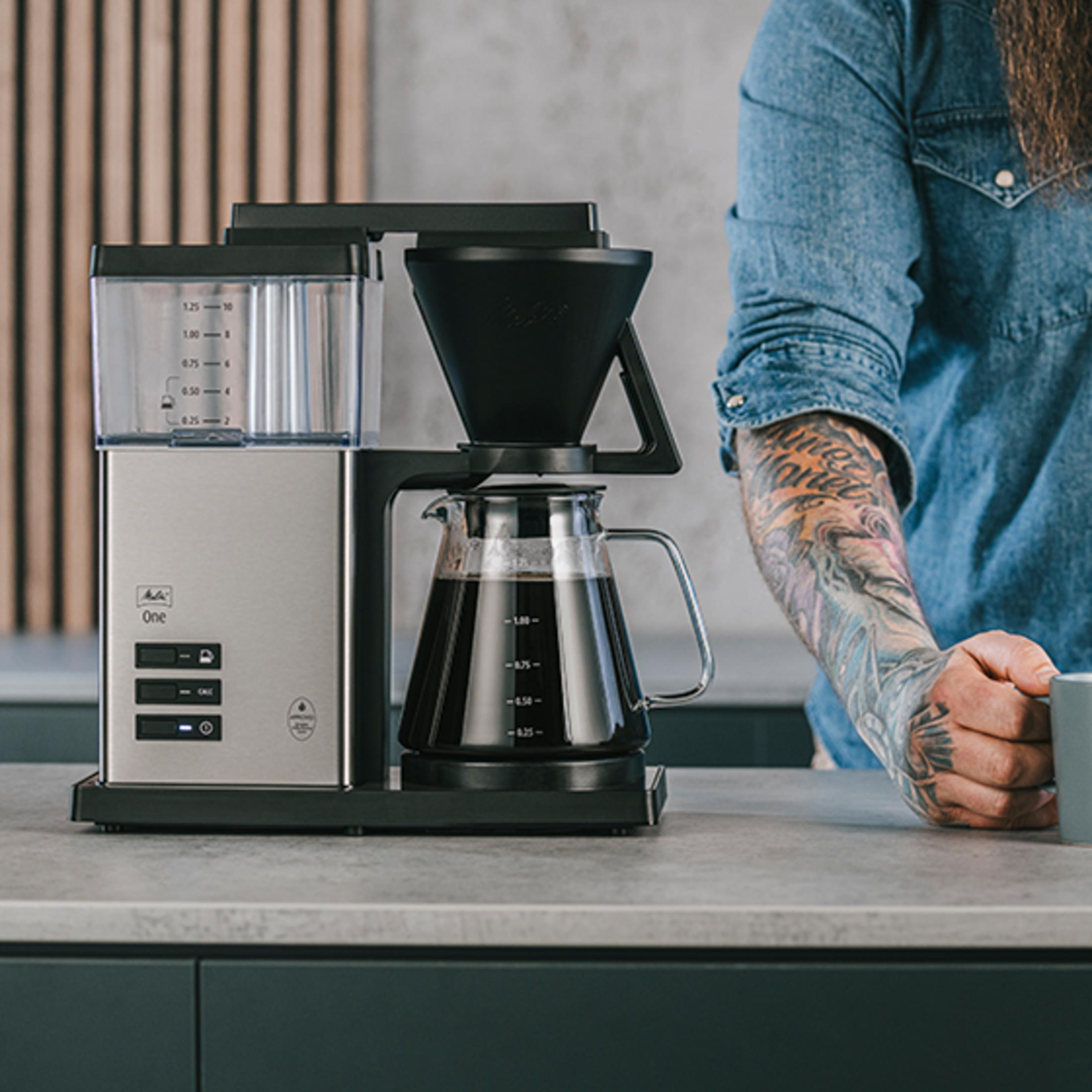 Une cafetière argentée avec une carafe, à côté d'une personne tatouée.