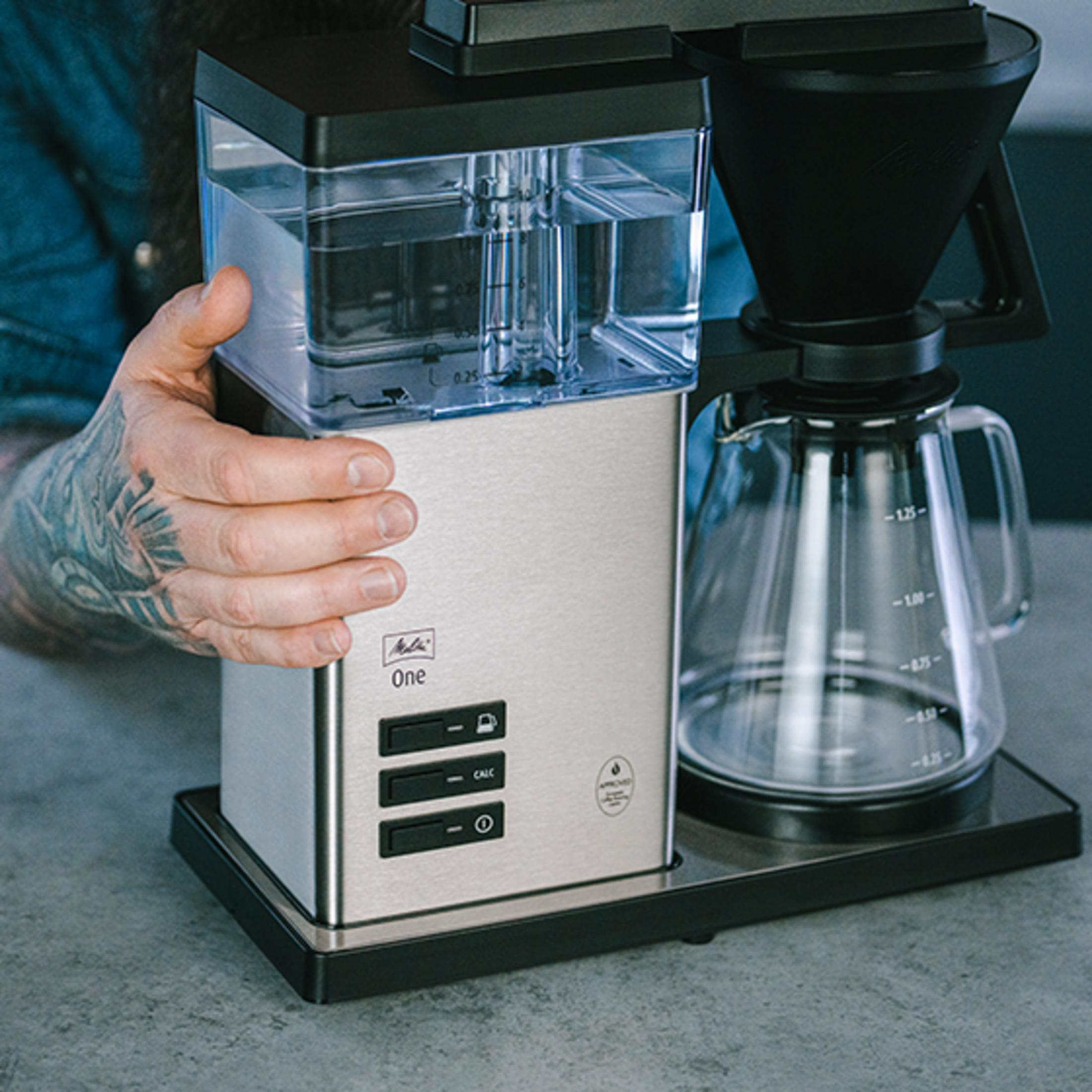 Une main tatouée remplit d'eau une cafetière avec carafe en verre.