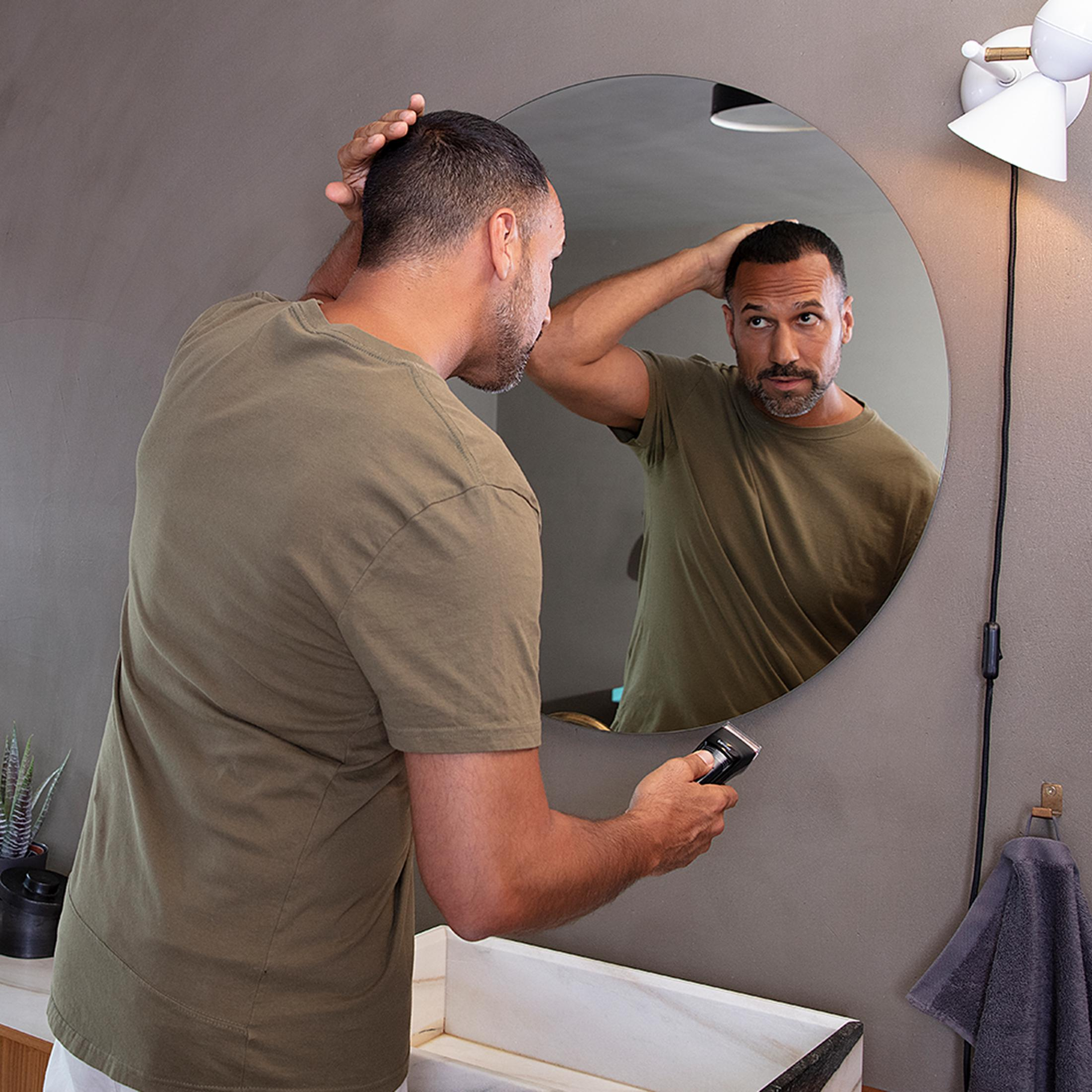 A man in an olive green t-shirt grooms himself in the mirror, holding a trimmer.