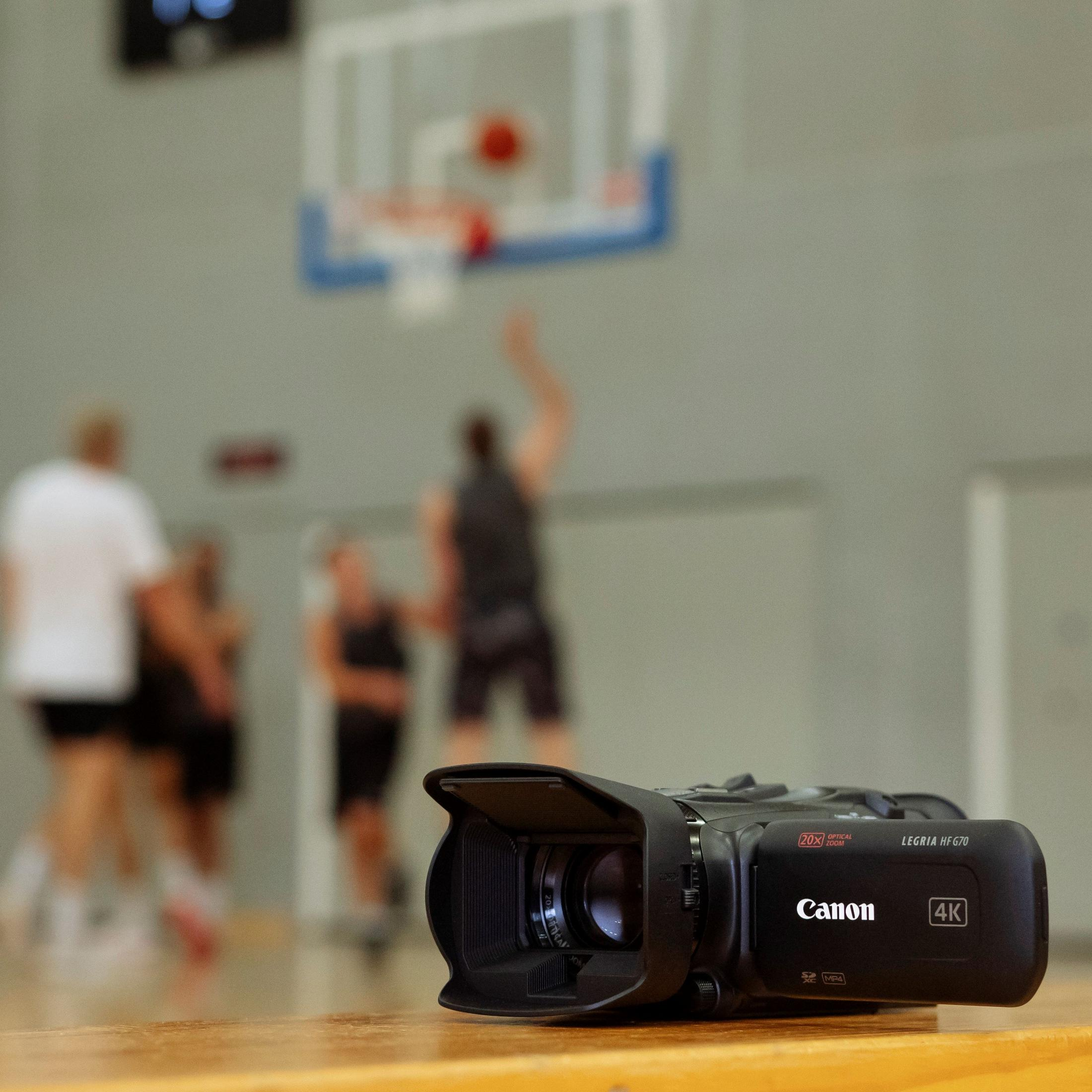 Una videocámara negra descansa sobre una mesa de madera con gente jugando al baloncesto detrás.