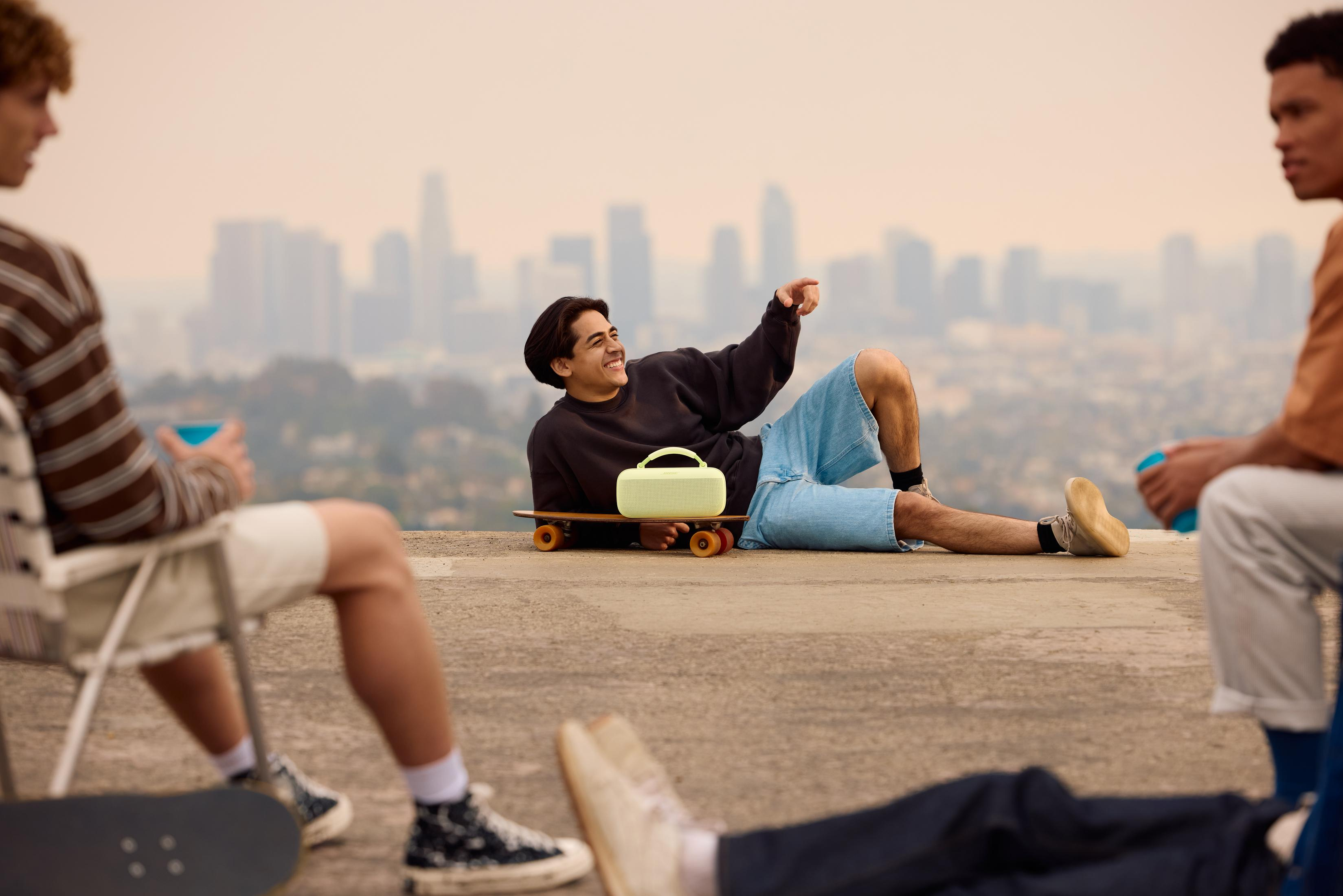 Mężczyzna leży na deskorolce, obok głośnik, w tle panorama miasta.