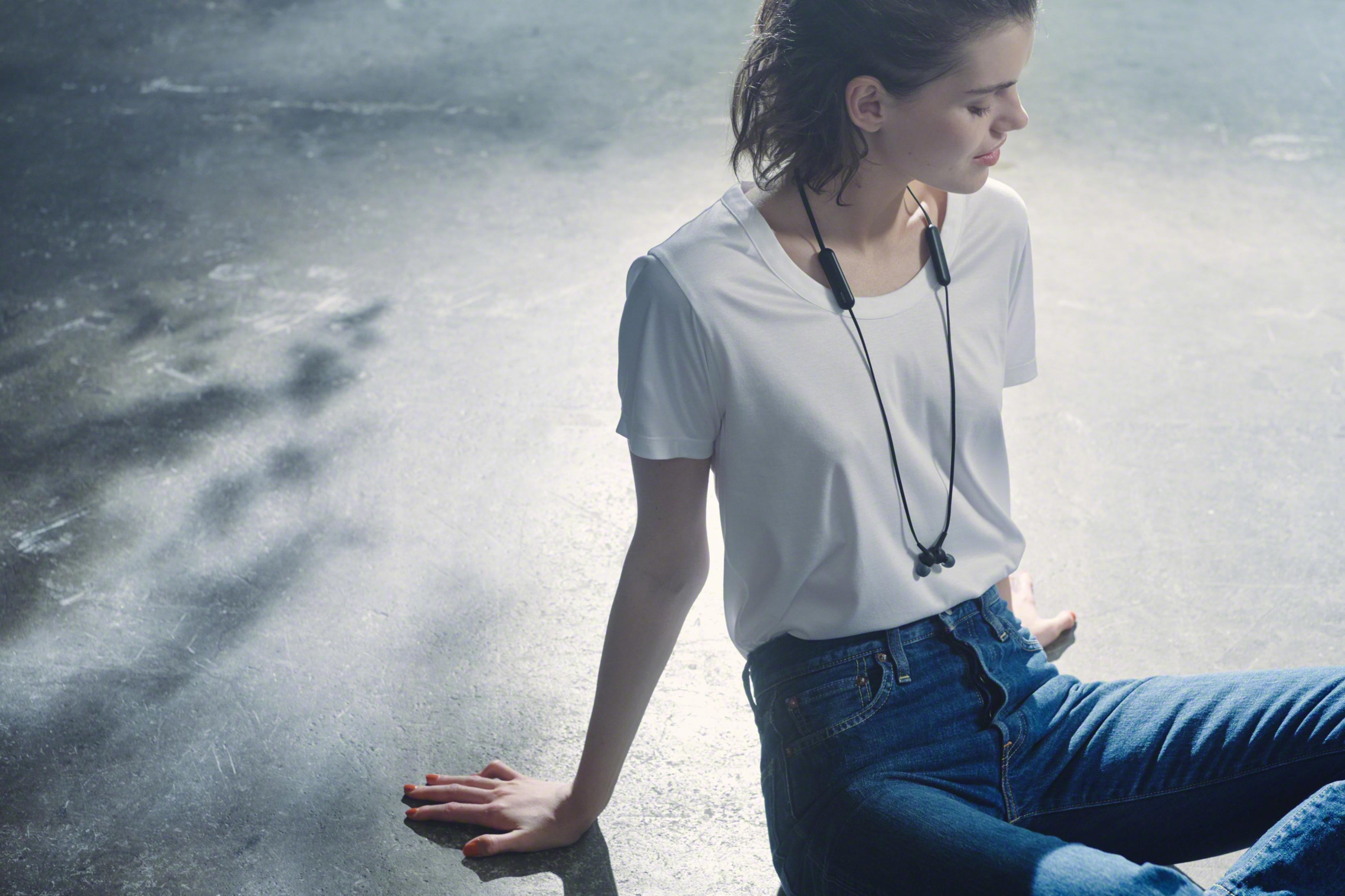 Młoda kobieta ze słuchawkami siedzi na szarej podłodze, ubrana w białą koszulkę i jeansy.