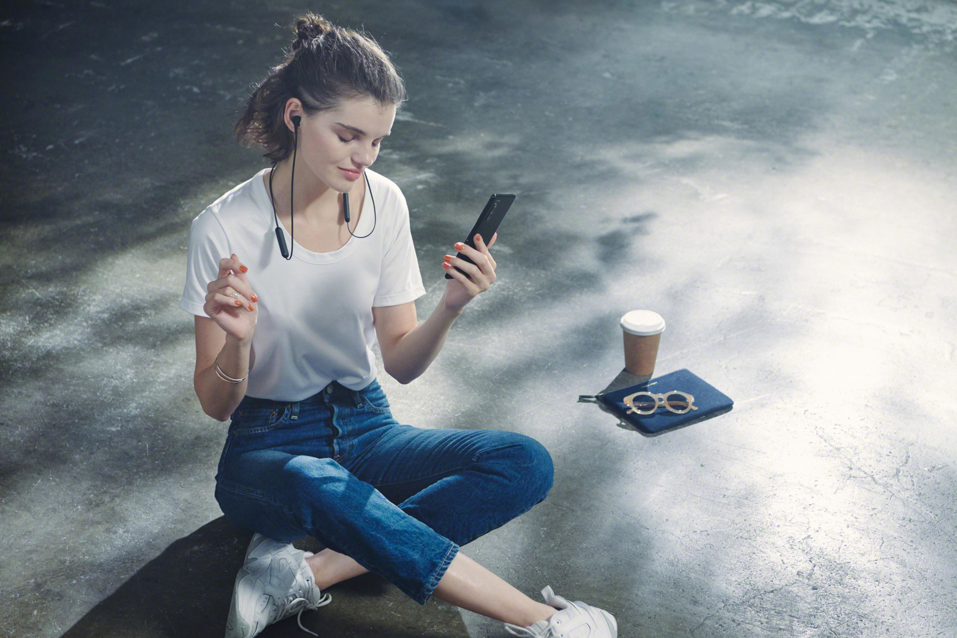 Kobieta ze słuchawkami siedzi i patrzy na telefon. Obok leży torba, okulary przeciwsłoneczne i kawa.