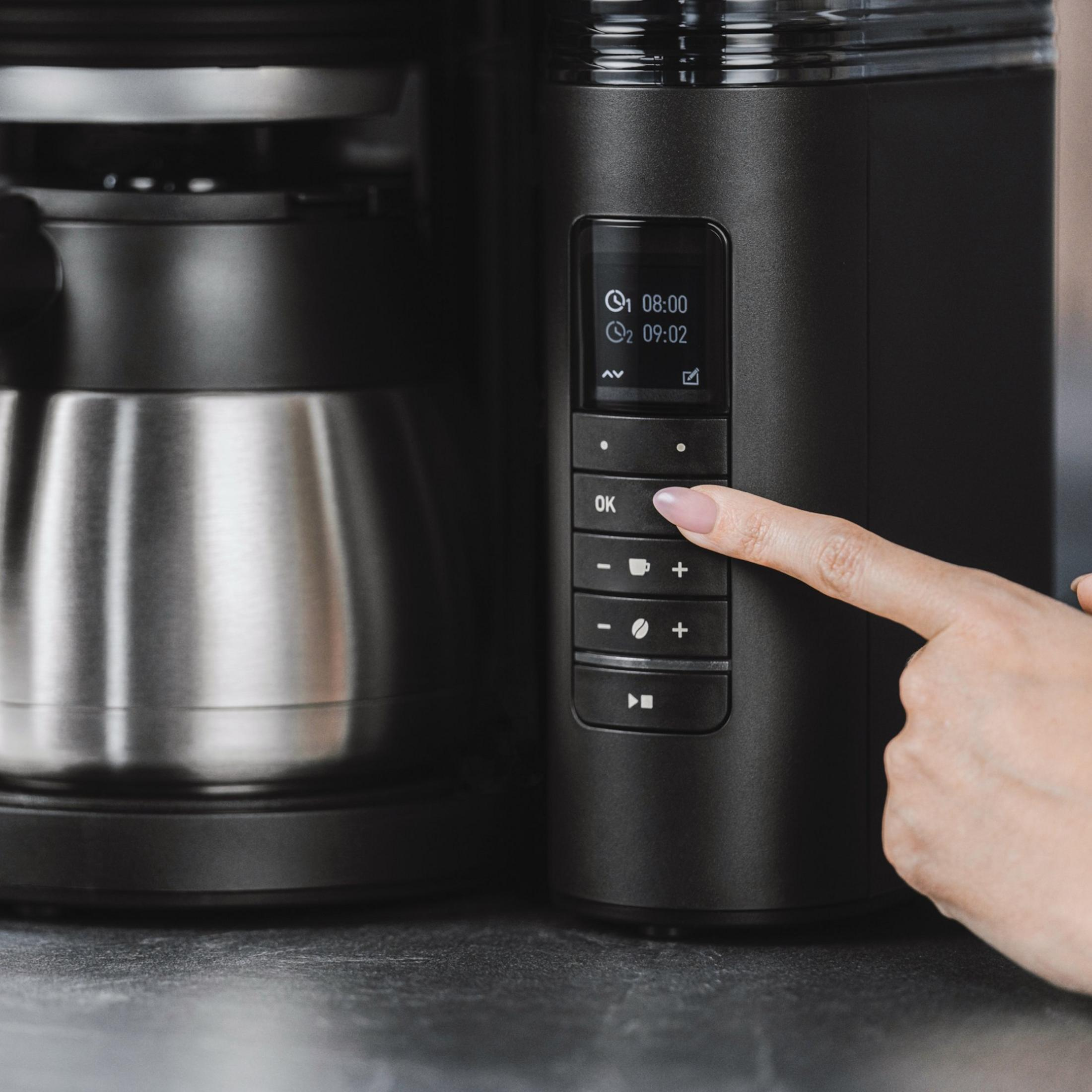 Dłoń naciska przycisk OK na czarnym ekspresie do kawy z cyfrowym wyświetlaczem.