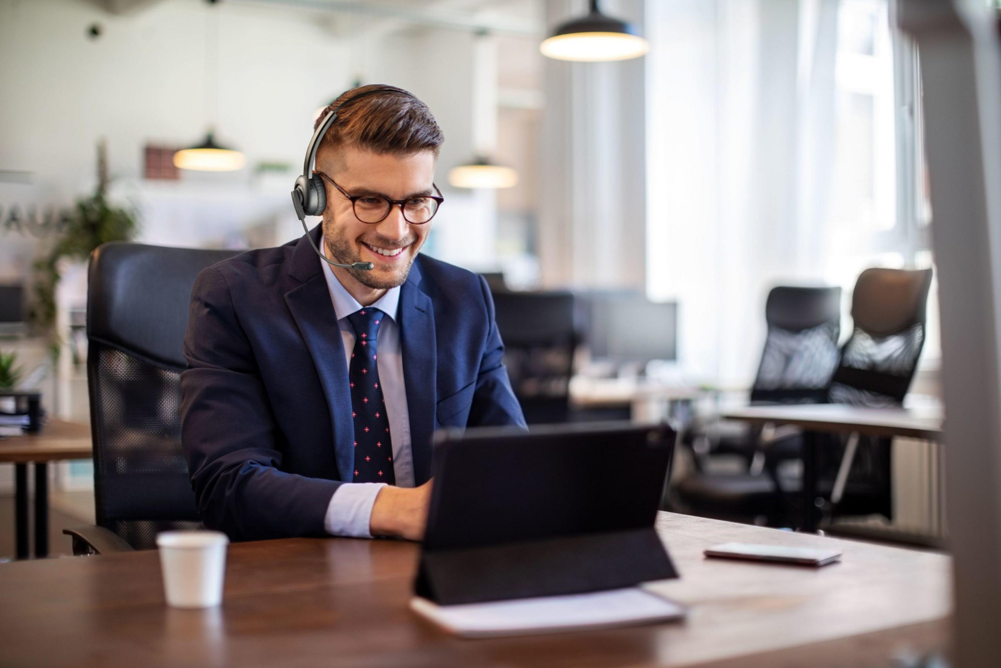 Ein Mann mit Brille und Headset sitzt am Schreibtisch und lächelt.