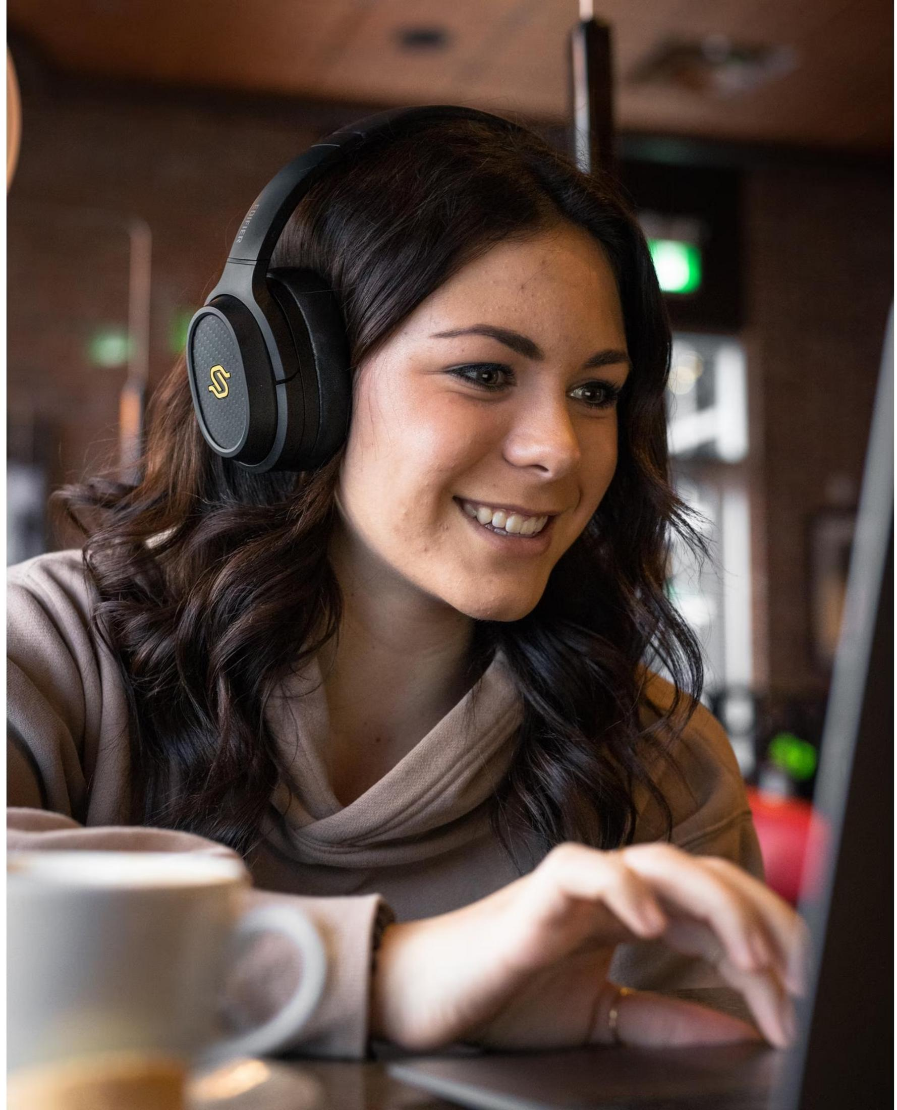 Una donna con cuffie lavora al laptop accanto a una tazza di caffè.