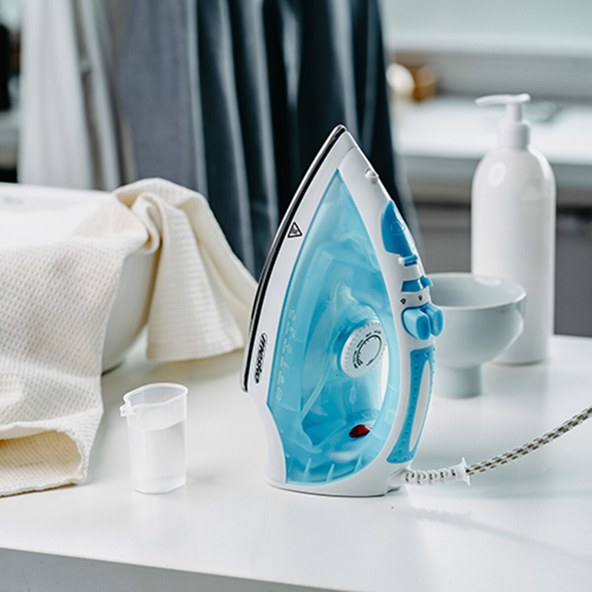 Ein Bügeleisen mit blauem Wassertank steht auf einem Tisch neben einem Messbecher mit Wasser.