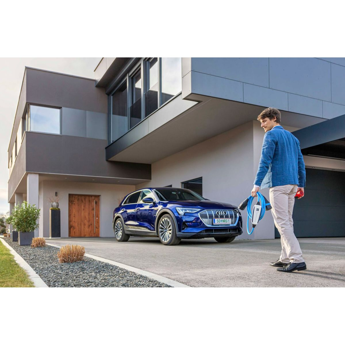 Mann mit Ladekabel steht neben blauem Elektroauto vor modernem Haus.
