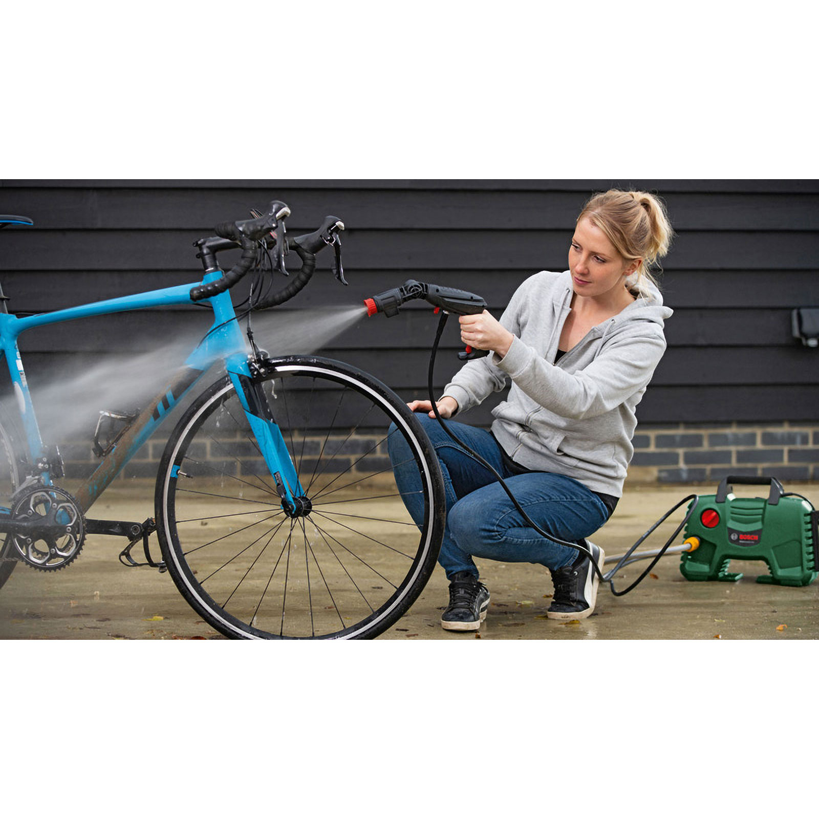 Een vrouw reinigt een blauwe fiets met een hogedrukreiniger voor een donkere houten muur.