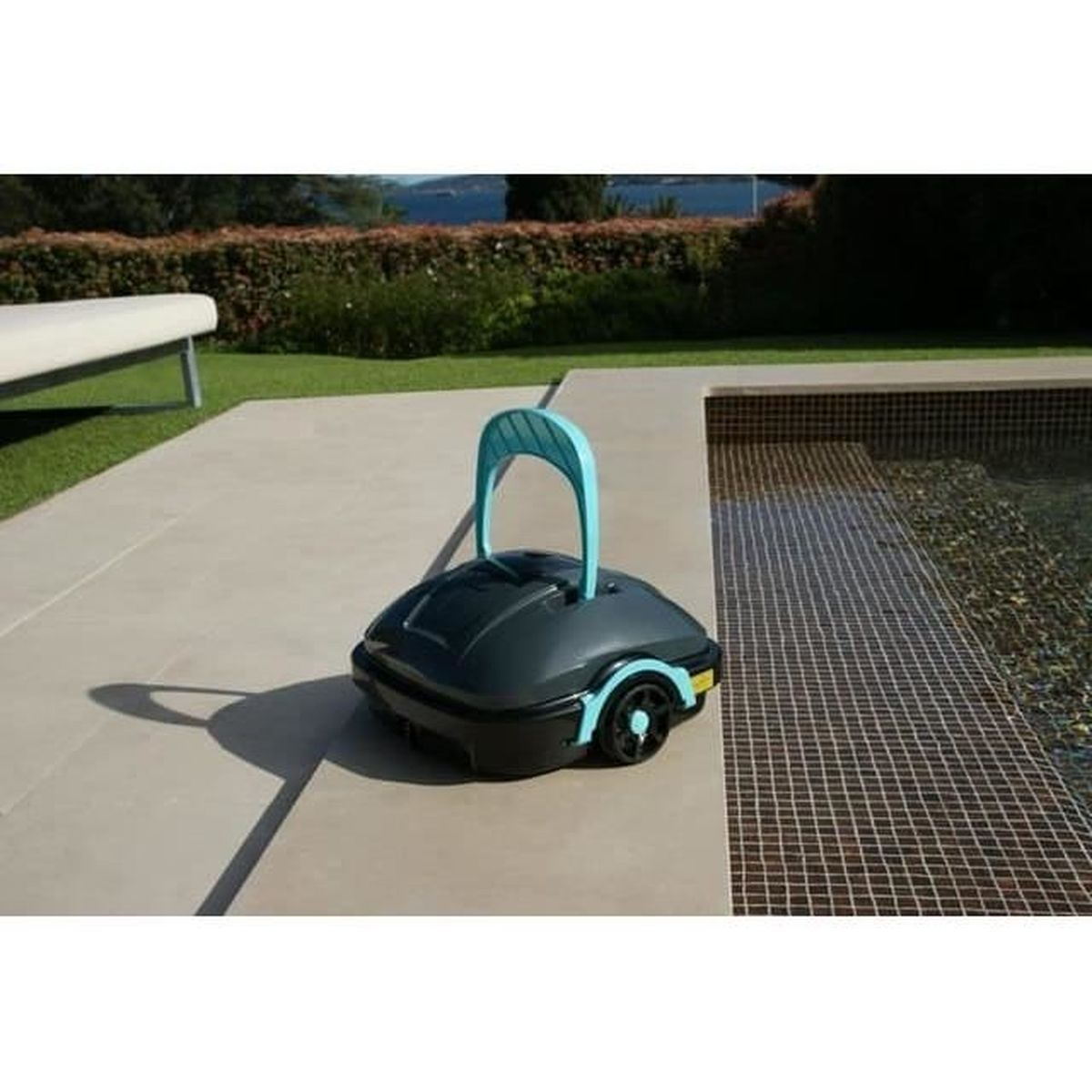Ein dunkelgrauer Poolreiniger mit türkisfarbenem Griff steht auf einer Steinplatte neben einem Pool.