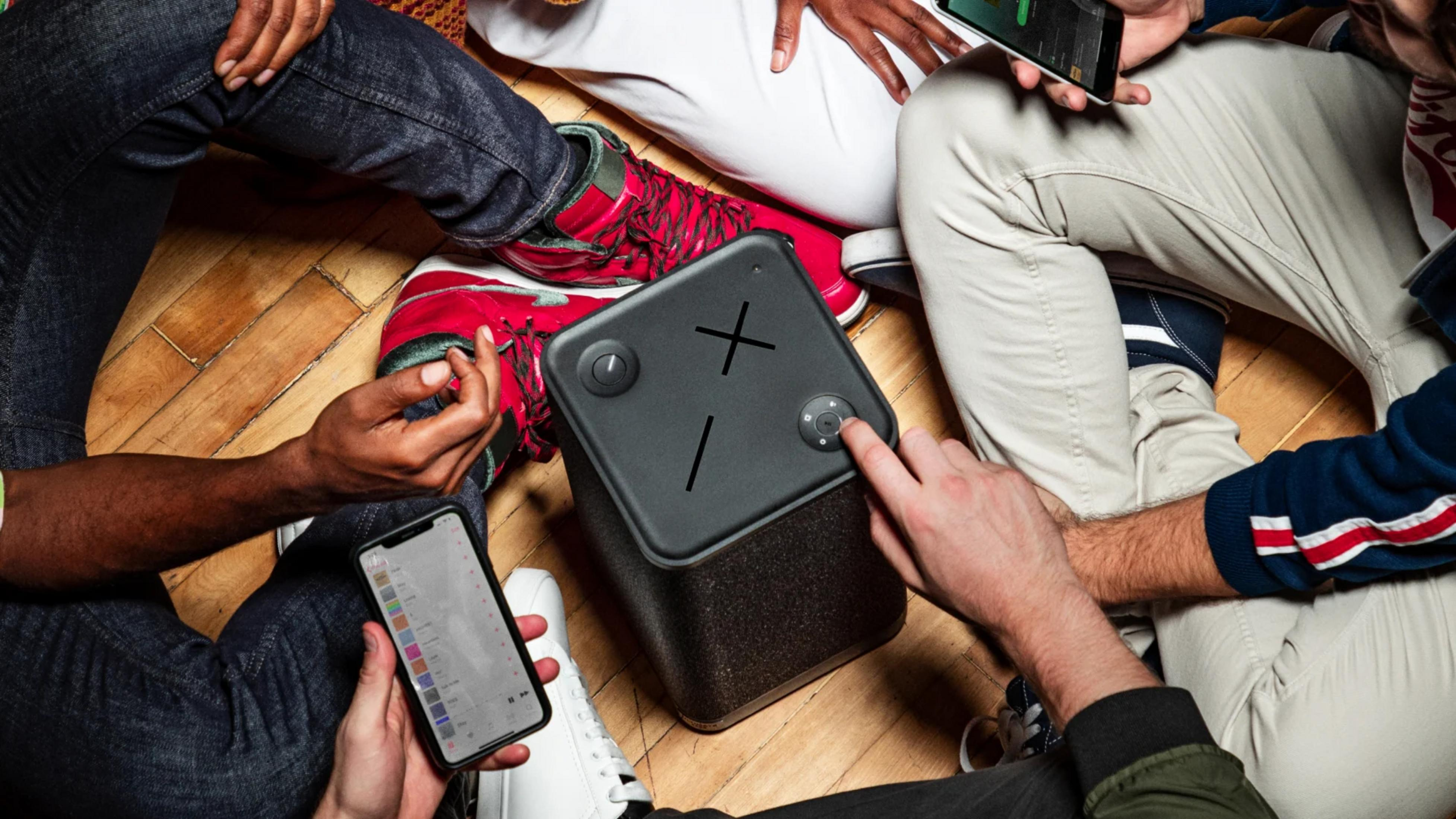 Ludzie siedzą na podłodze wokół głośnika i smartfonów.