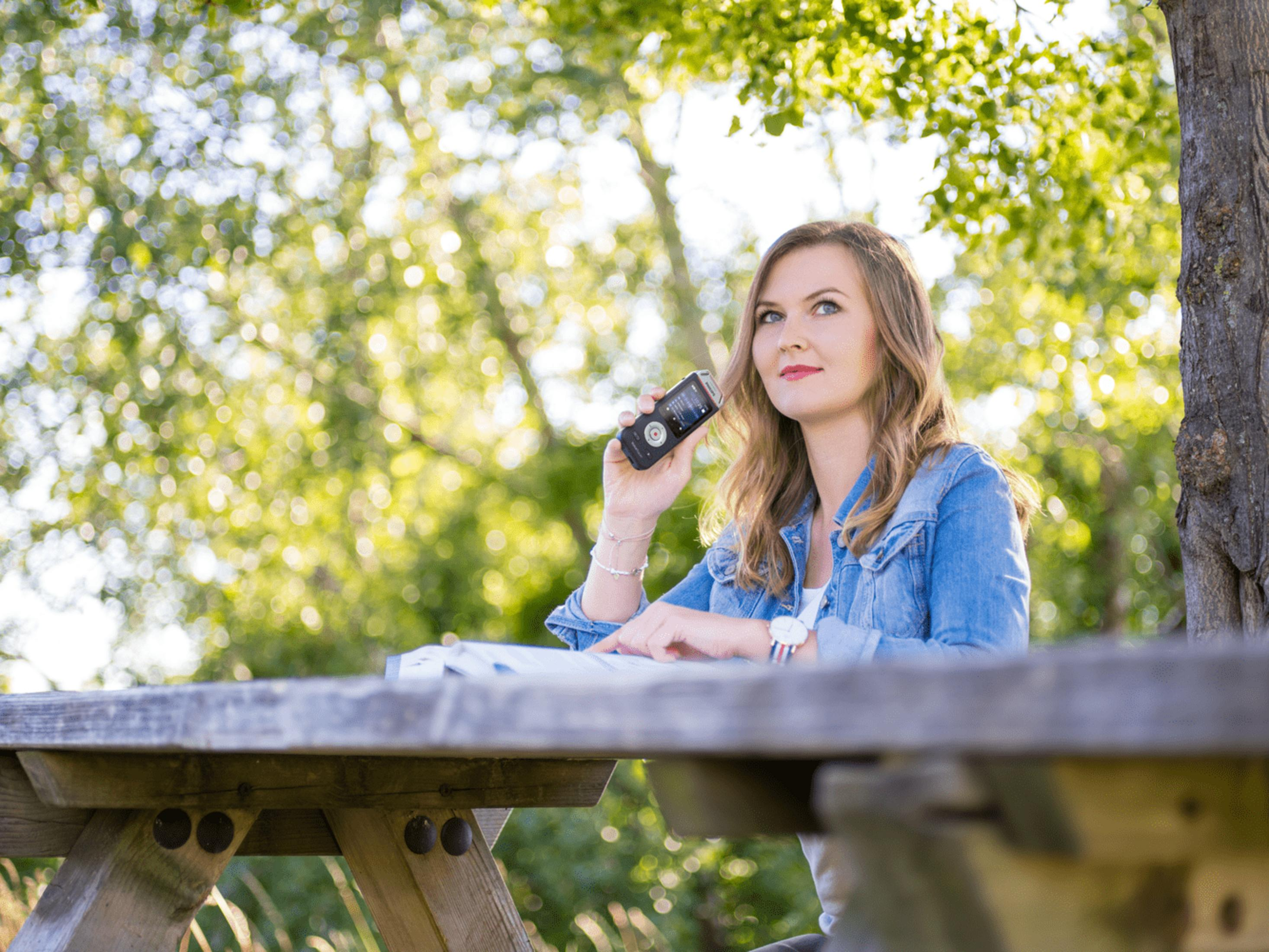 Kobieta siedzi przy drewnianym stole na zewnątrz, trzymając telefon komórkowy.