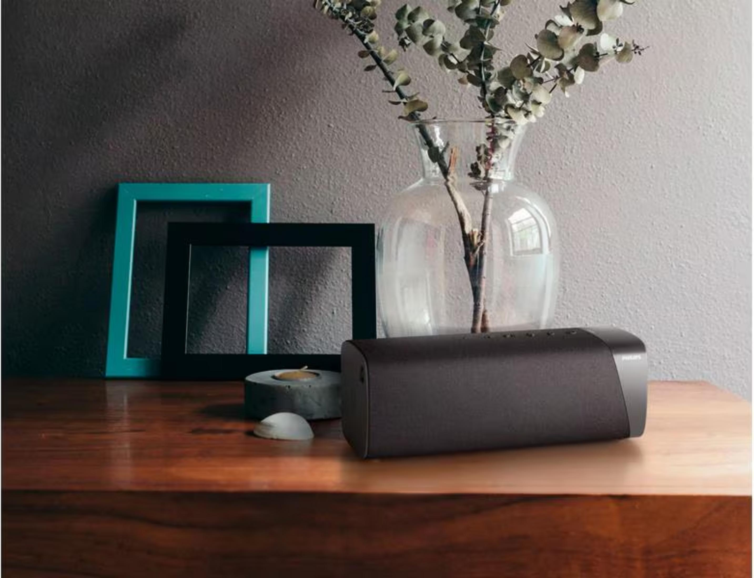 Ciemny głośnik leży na drewnianym stole obok ramek na zdjęcia i wazonu.