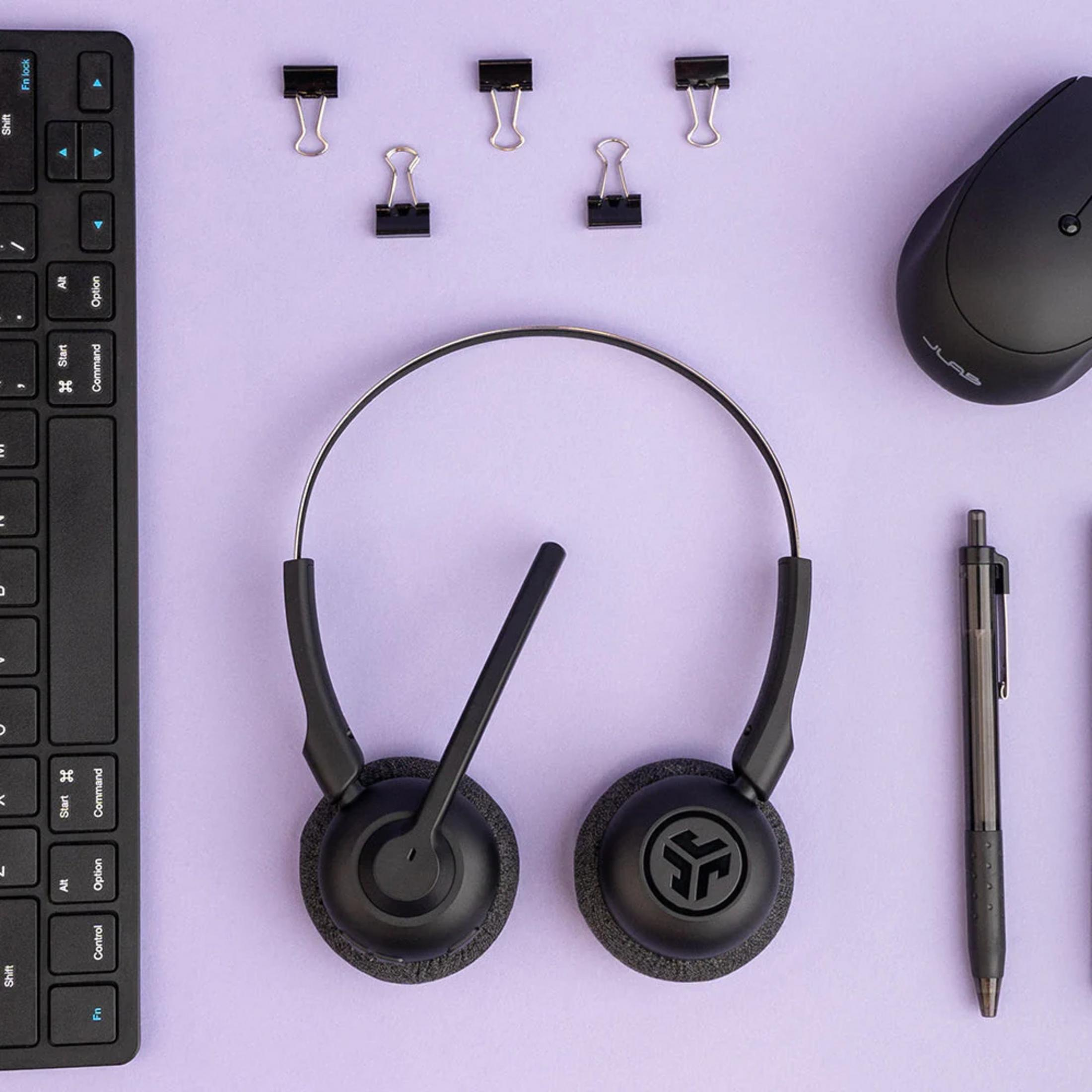 Czarne słuchawki, mysz, klawiatura, długopis i spinacze na fioletowym tle.