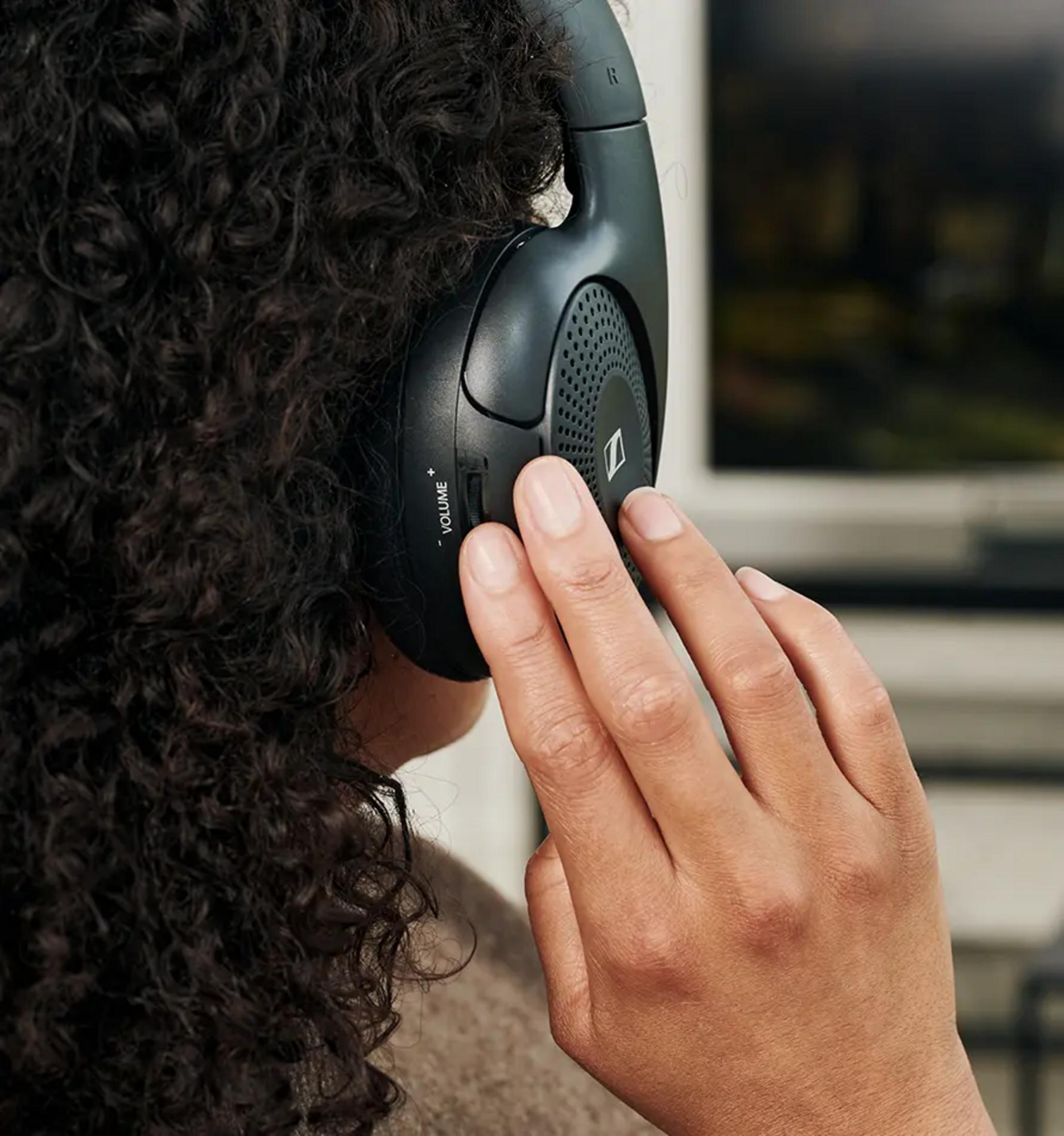 A person with curly hair wears black headphones, a hand adjusts the volume.