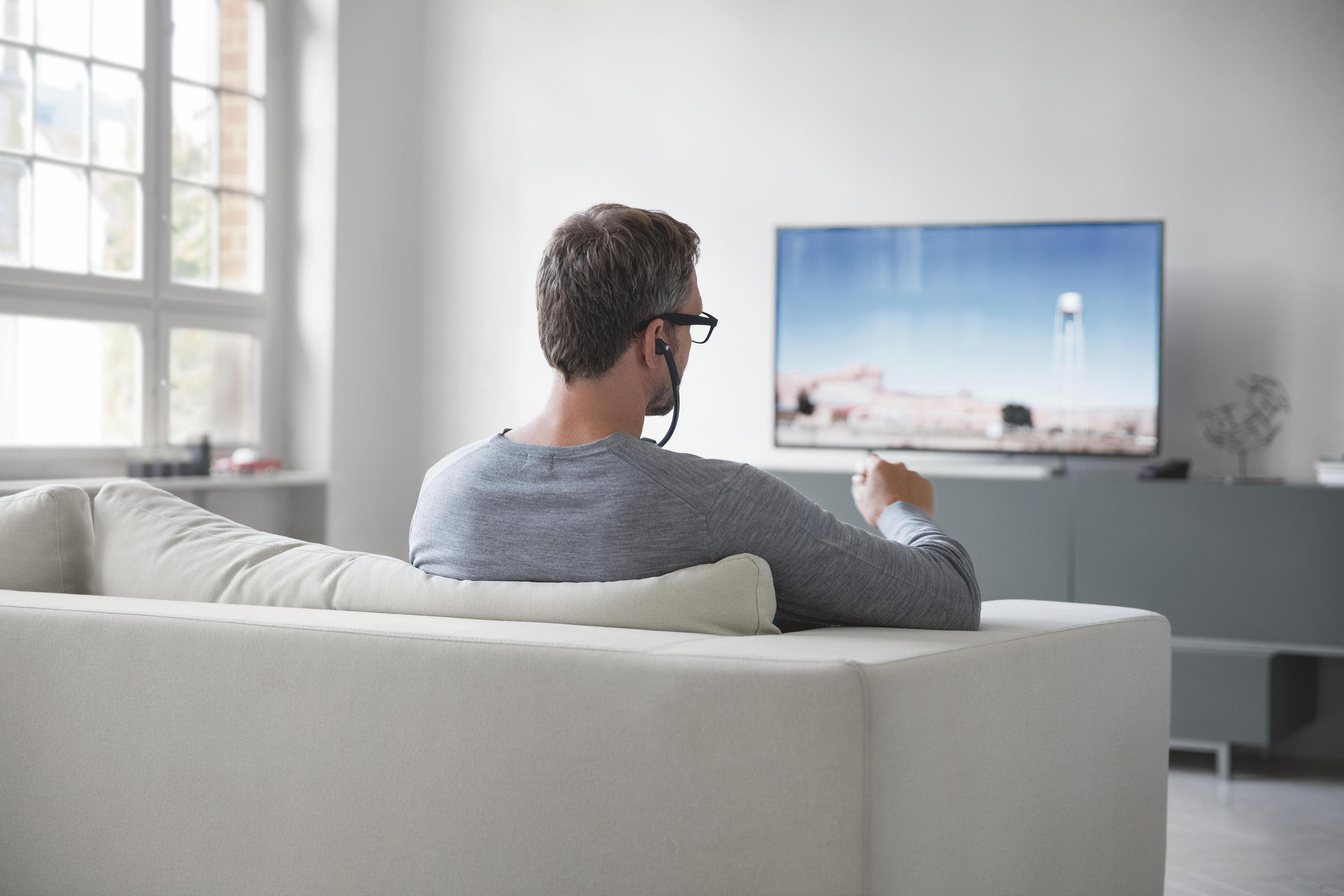 Mann sitzt auf Sofa und schaut fern. Graues Hemd, Brille, Kopfhörer.