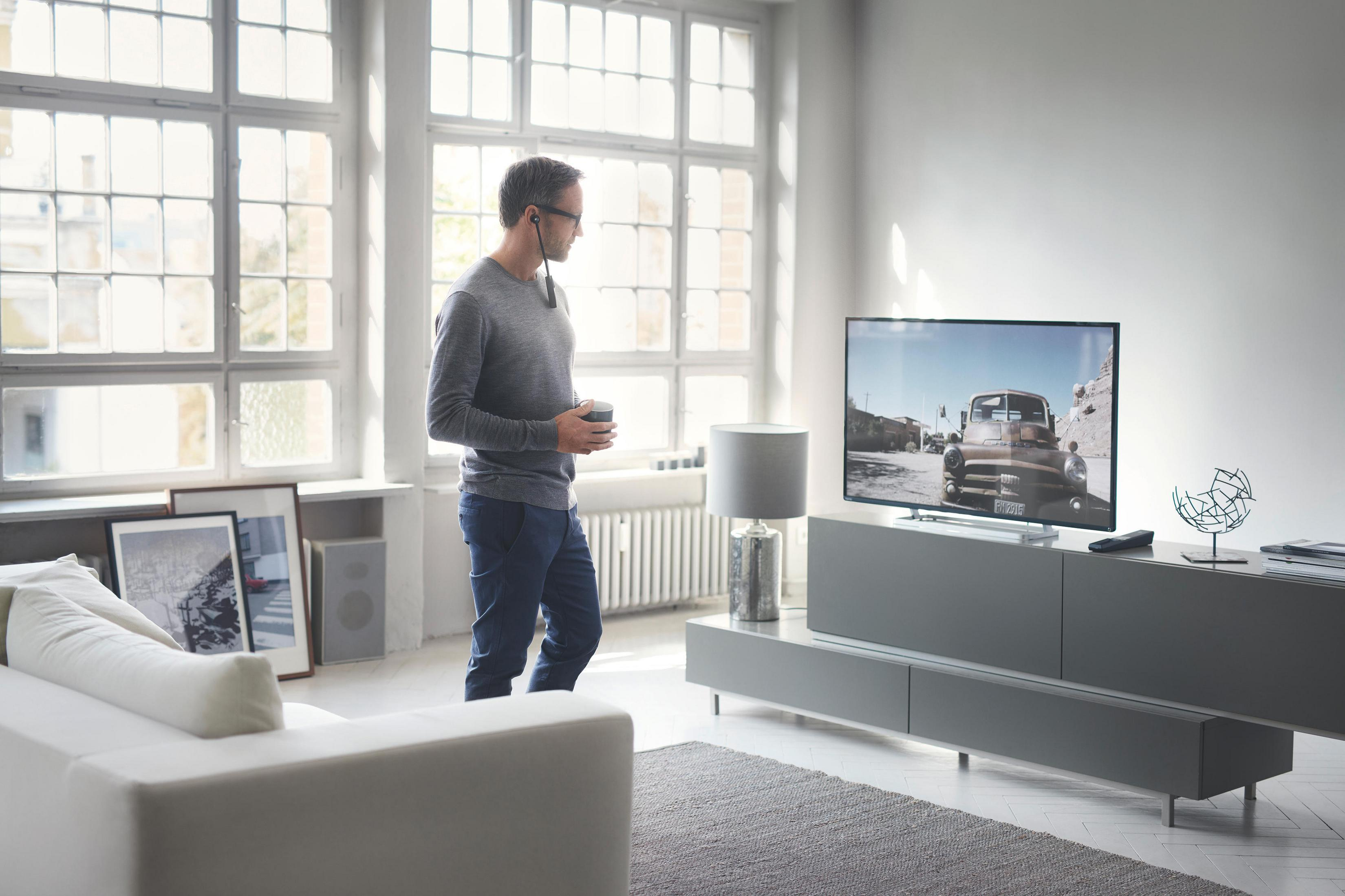 Mann im Wohnzimmer schaut Fernsehen, hält eine Tasse, Fenster im Hintergrund.