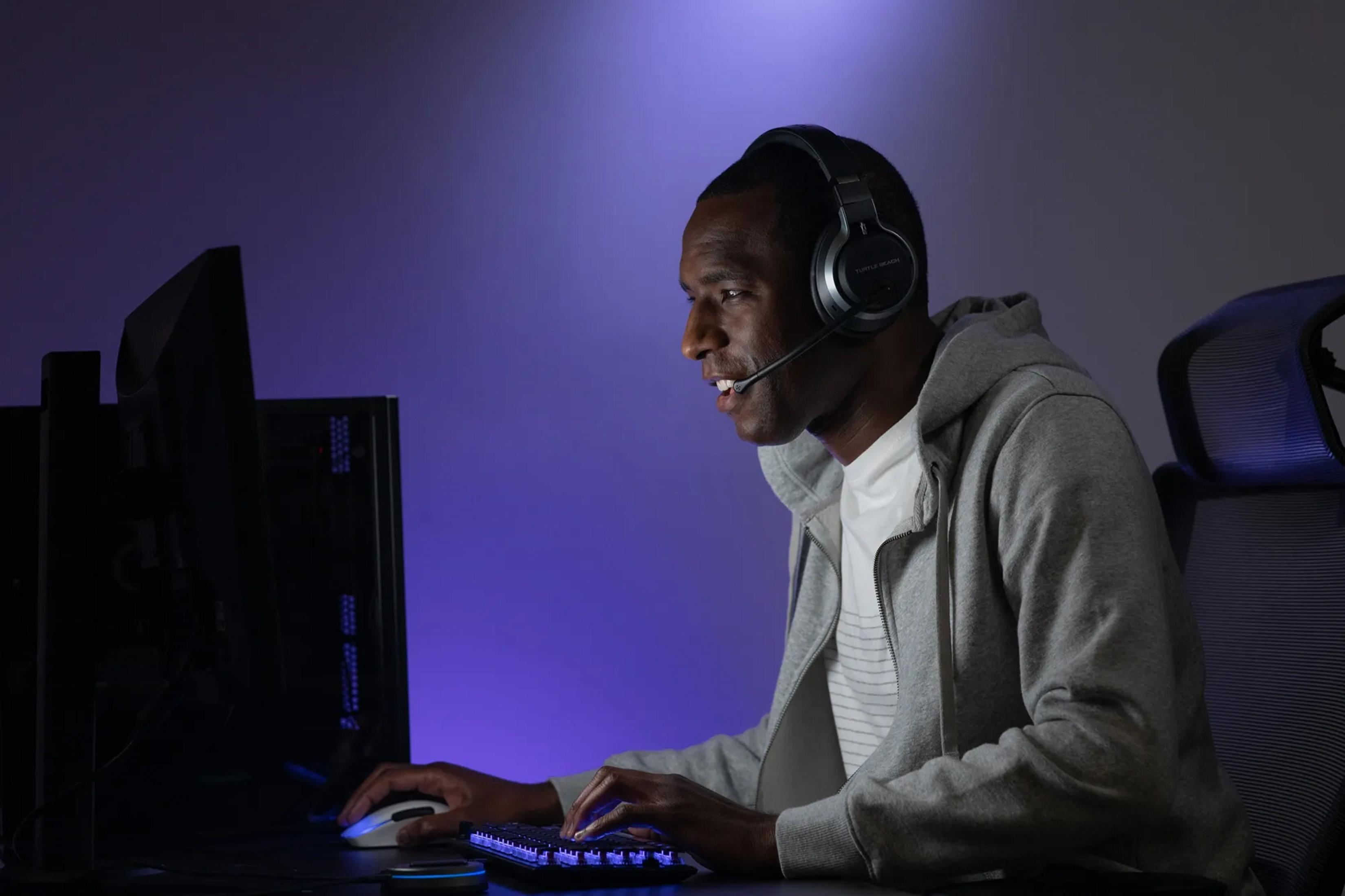 Mężczyzna w słuchawkach gra na komputerze, oświetlony fioletowym światłem.